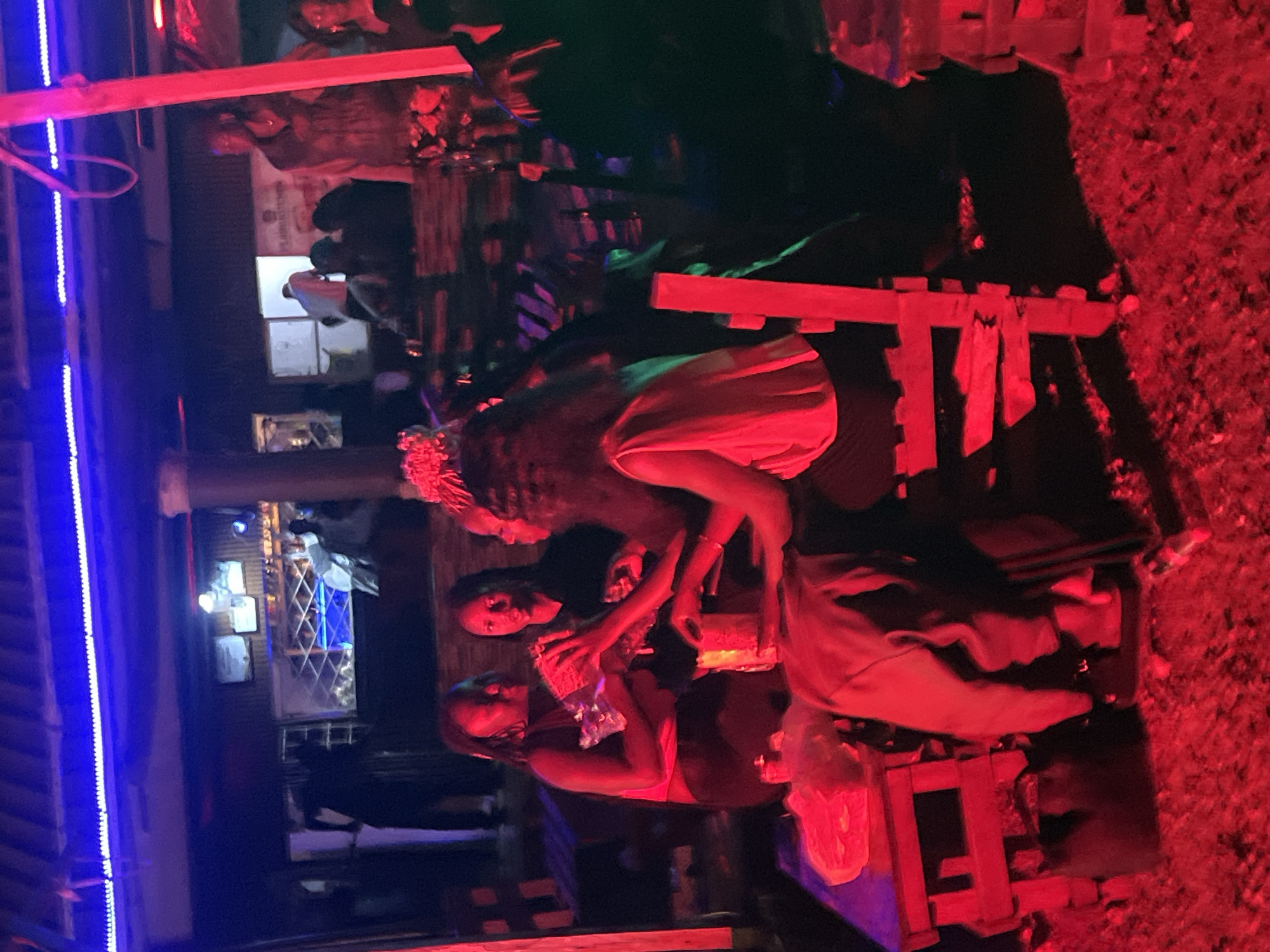 In Durumi, Nigeria, a dimly lit outdoor establishment features a gathering of individuals seated at rustic wooden tables under a corrugated metal roof illuminated by vibrant blue and red lights. Three women are the primary focus, engaged in conversation and interacting with a packaged item on the table. The woman on the left, with dreadlocks, wears a dark sleeveless top and holds the package. Next to her, another woman in a black top looks towards the package. To her right, a third woman with a tiara-like hair accessory, wearing a light-colored, short-sleeved top and dark bottoms, leans forward, observing the interaction. The environment appears to be a casual outdoor bar or social area, with other patrons faintly visible in the background. The ground is covered in gravel or small stones, reflecting the ambient red light.