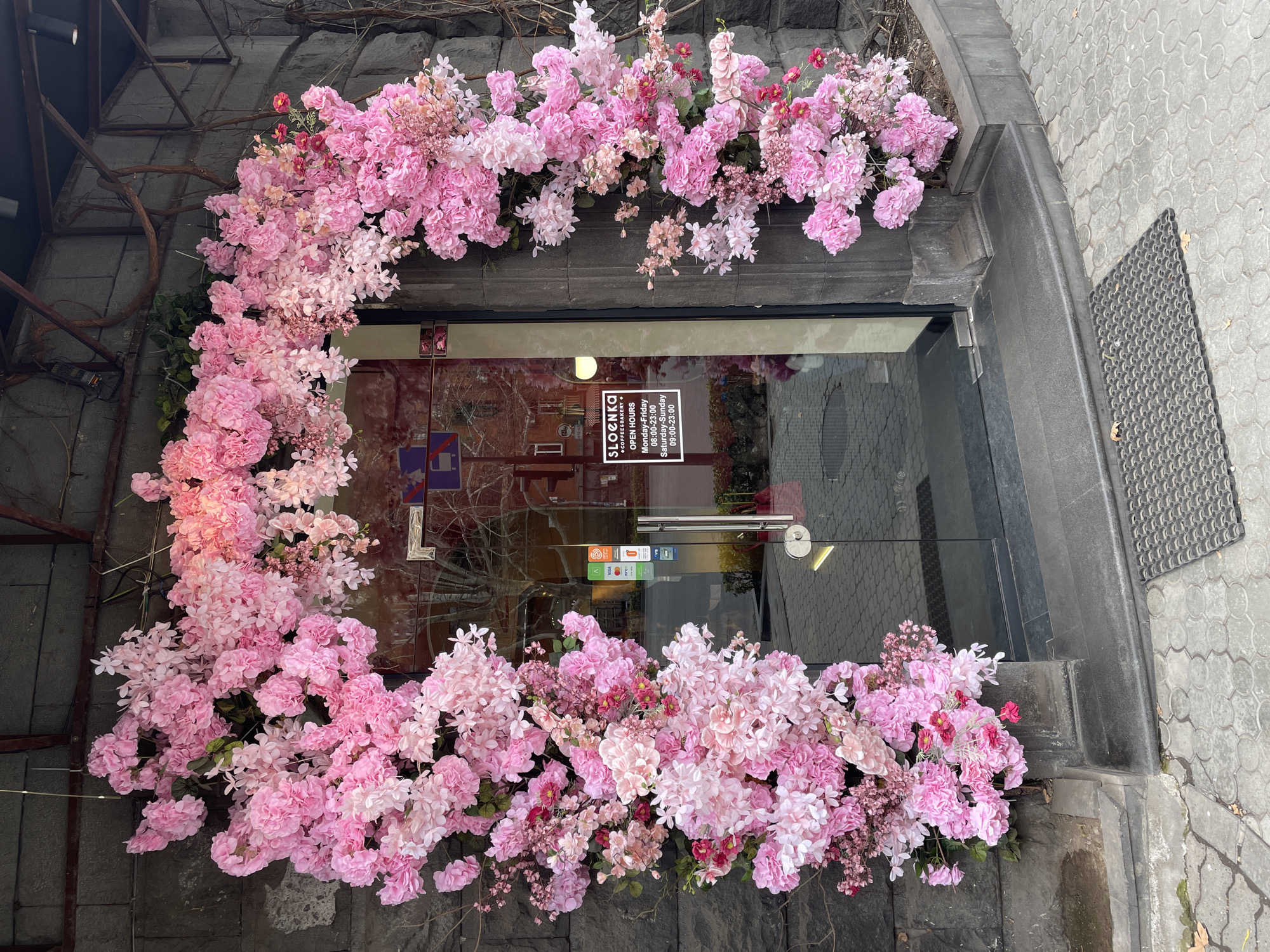 The image shows the entrance of "Sloenka Coffee & Bakery" in Yerevan, Armenia. The doorway is decorated with an abundance of pink flowers, creating a floral frame around the glass door. The building's exterior is made of stone, with a gray stone pavement leading to the entrance and a rubber mat on the right side. A sign on the door indicates the bakery's name and hours of operation: Monday-Friday from 08:00 to 23:00 and Saturday-Sunday from 09:00 to 23:00. The reflection in the glass shows part of the interior and a blurred view of the street.