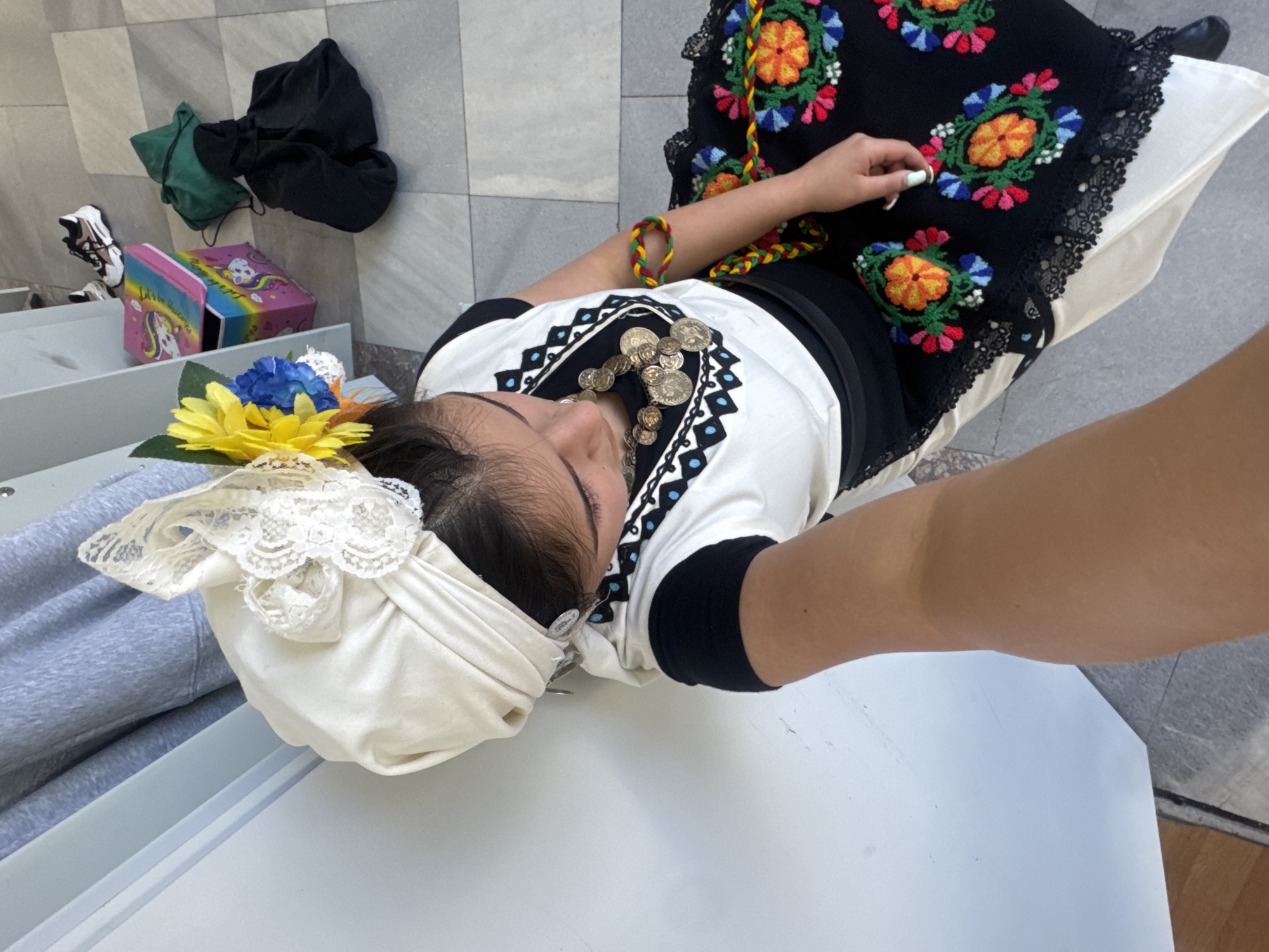 A high-angle shot captures a person wearing traditional attire, possibly engaged in a cultural event or performance. The individual is adorned with a white head covering featuring lace and a yellow flower with blue accents. They are also wearing a white top with black trim, embellished with metal coin-like adornments. The traditional dress has a black embroidered skirt with vibrant floral designs and a lace trim. The person is wearing a multicolored rope-like bracelet.
In the background, there is a grey wall with a black bag and a green bag, along with what appears to be a shoe and a colorful box. The setting seems to be indoors, with a table or counter in the foreground.