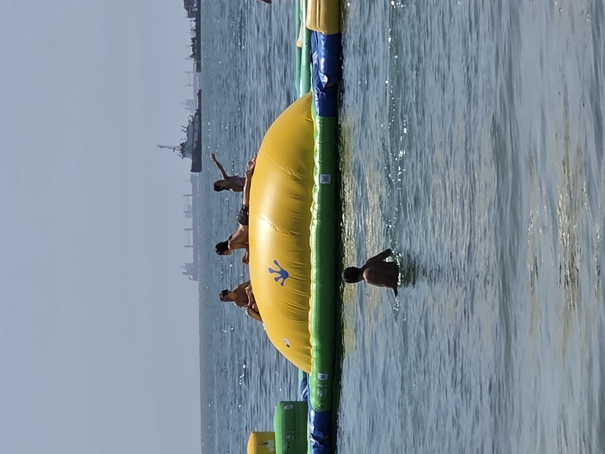 The image captures a sunny day at an outdoor water park in Long Beach, USA. A group of young people are enjoying a large inflatable water obstacle course in the ocean. The main subject is a vibrant yellow inflatable structure, part of the obstacle course, with a blue frog decal. Several individuals, appearing to be teenagers or young adults, are either on the inflatable or in the water nearby. Some are perched on the edge of the yellow structure, seemingly preparing to jump, while others are already in the water. One person is clearly visible in the water to the right of the inflatable, facing away from the camera. The water is a clear blue, with sunlight glinting off the surface. In the background, a hazy sky suggests a warm and clear day. The silhouette of a ship is visible in the distance on the left, hinting at the proximity of the port or coastline. The overall mood is one of active fun and recreation. There is no visible text in the image.
