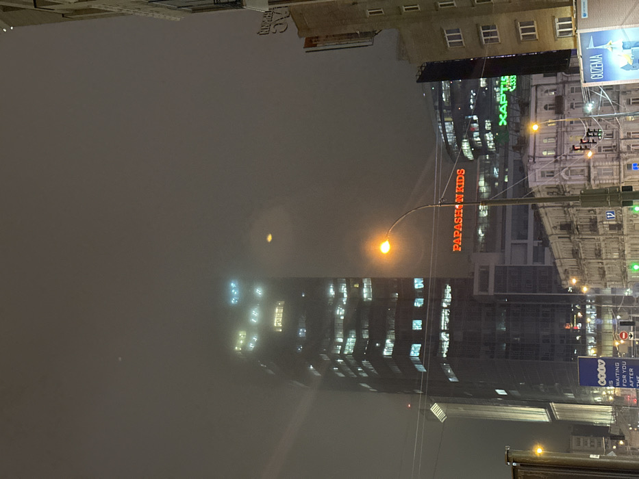 A foggy night scene features illuminated buildings. "PAPASHON KIDS" is displayed vertically in red lettering on one building. A curved building with green neon "XAPTIC" is visible, along with a traditional building with lit windows. A streetlamp glows in the foreground, and traffic lights are visible in the distance. Street signs and buildings recede into the fog.