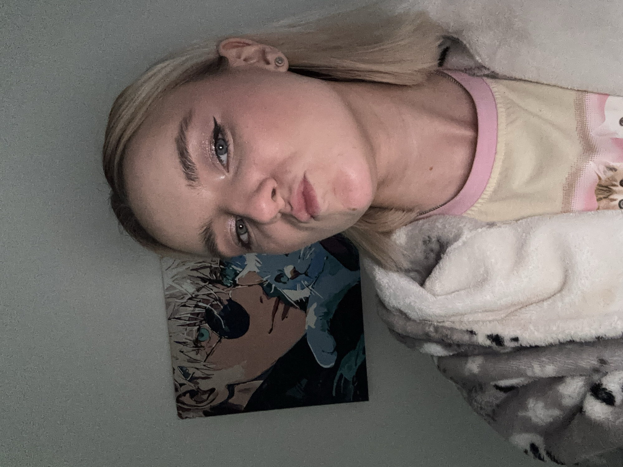 A young woman with blonde hair and blue eyes poses for a selfie. She has eyeliner and is pouting. She is wearing a light-colored shirt with a pink trim. She is wrapped in a white and gray blanket. On the wall behind her is a poster featuring an anime character. The location is not identifiable.