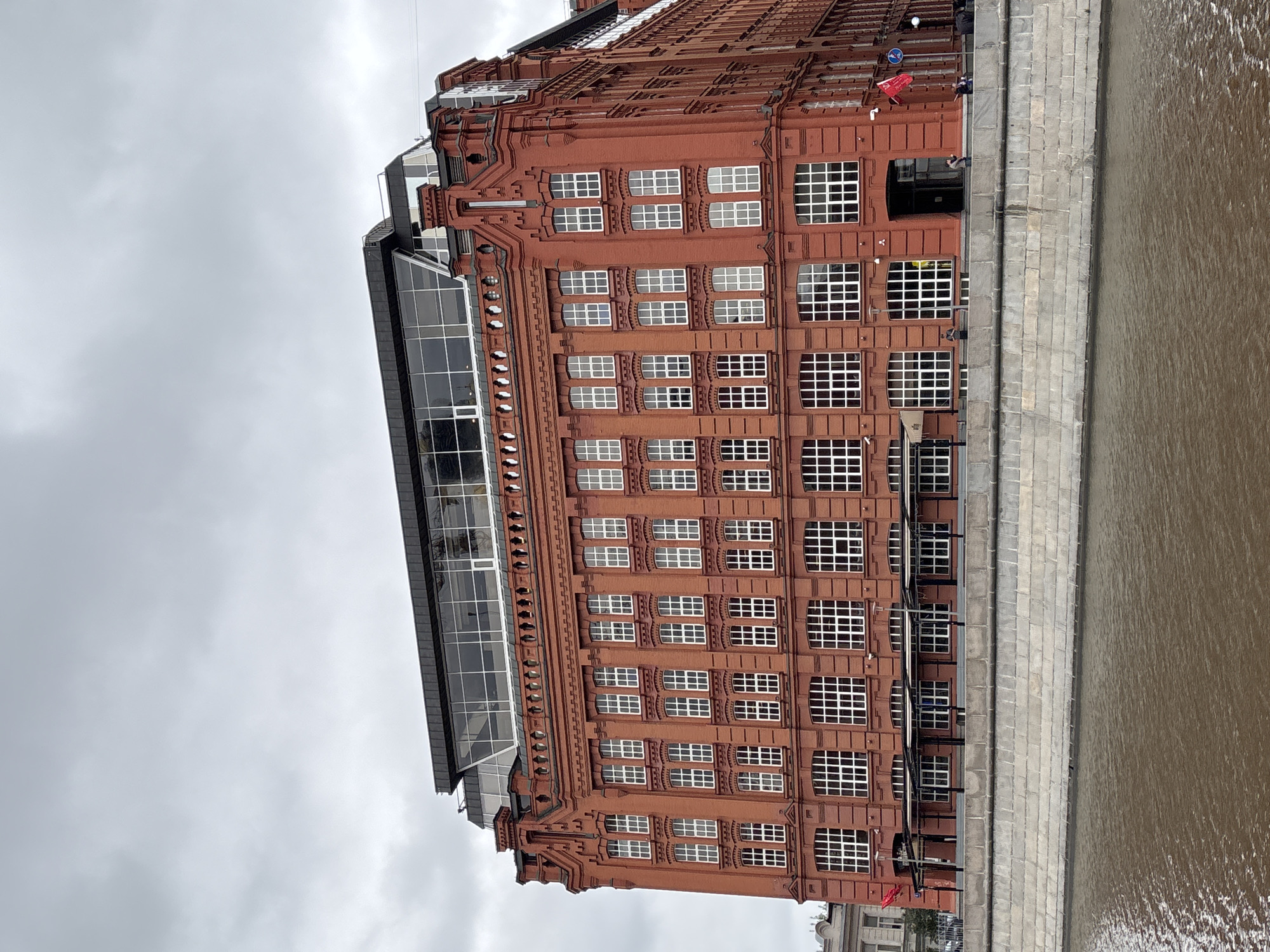 A vertical, slightly angled shot shows a red brick building, likely a factory or industrial structure converted to another use, next to a river. The building is tall, several stories high, with a large number of square windows with white frames. The facade is decorated with intricate brickwork. A modern glass structure appears to be an addition or renovation to the original building. The river is a muddy brown color and is bordered by a stone embankment. The sky is overcast, suggesting a cloudy day. Several people are visible near the edge of the building closest to the river. Based on the architecture, the image is most likely taken in Moscow, Russia.