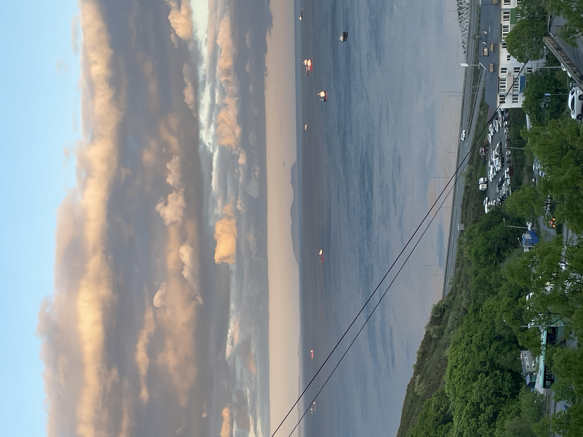 The image depicts a view from an elevated position, potentially a high-rise building, overlooking a cityscape, the sea, and the sky. The sea contains several ships. The sky contains multicolored clouds. The cityscape is on the right side and has a parking lot with white cars, buildings, a road, and green trees.
Location: Likely Vladivostok, Russia, based on the visual characteristics of the architecture, environment, and coastline.