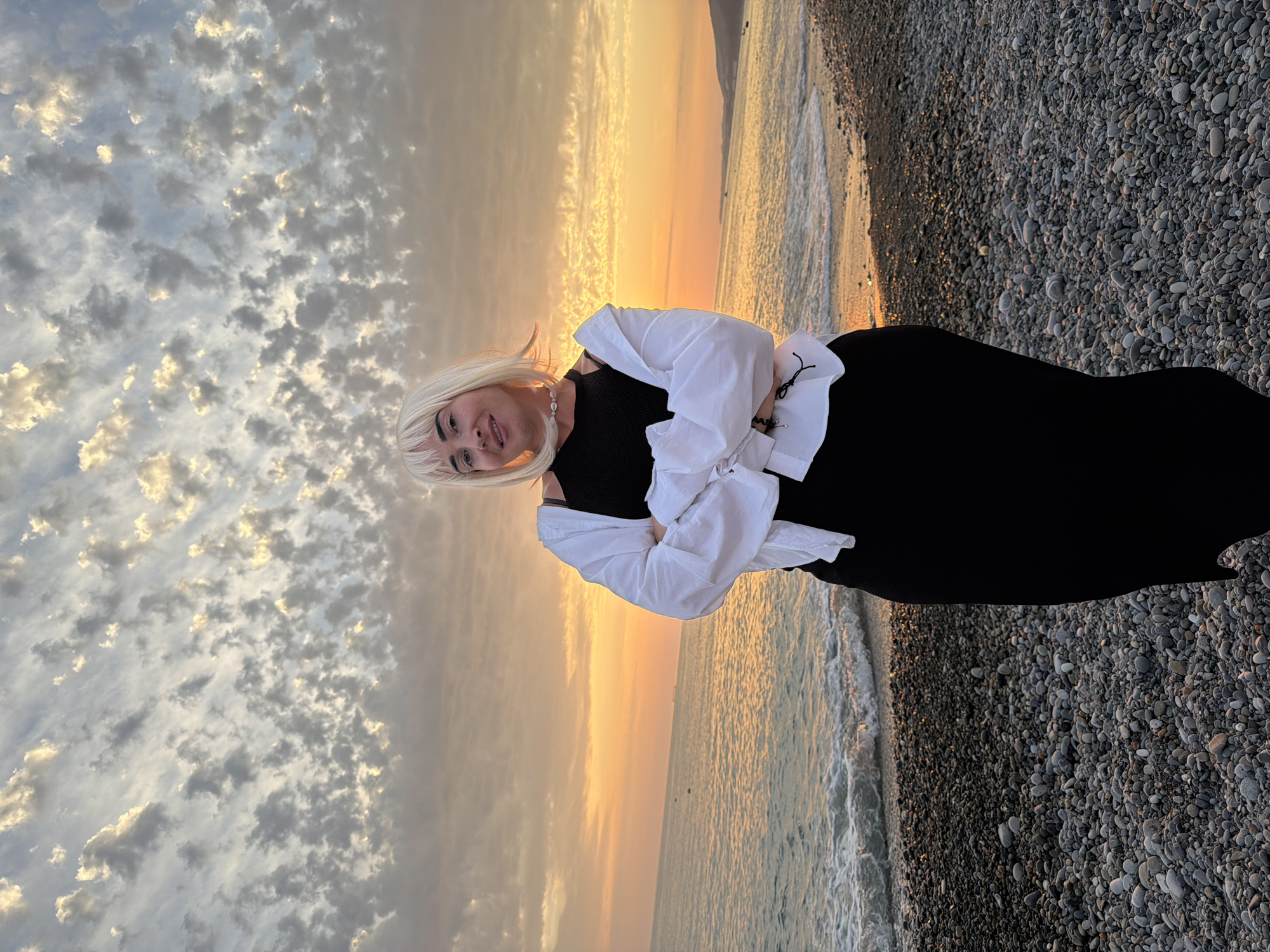 A full-body, eye-level, medium shot shows a woman standing on a pebble beach with her arms crossed in front of her. She has platinum blonde hair with bangs and is wearing a black tank top with a white open button-up shirt over it. She has a necklace with small white beads. The background shows the ocean with small waves, a distant shoreline, and a dramatic sunset with scattered clouds. The overall lighting suggests it is late evening.