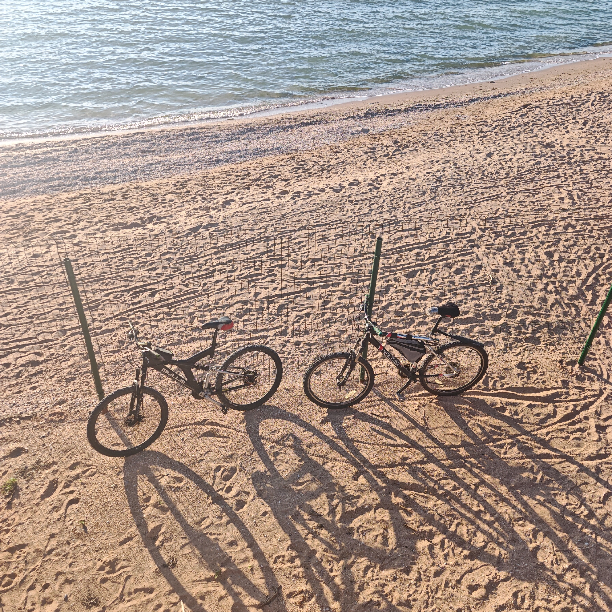 Two bicycles are parked on a sandy beach in the city of Shchelkino, Ukraine. The bicycles are positioned near a wire fence, with their shadows stretching across the sand. The sea is visible in the background, with calm waves lapping at the shore. The sand is imprinted with footprints and tire tracks, indicating recent activity. The lighting suggests it is either early morning or late afternoon.