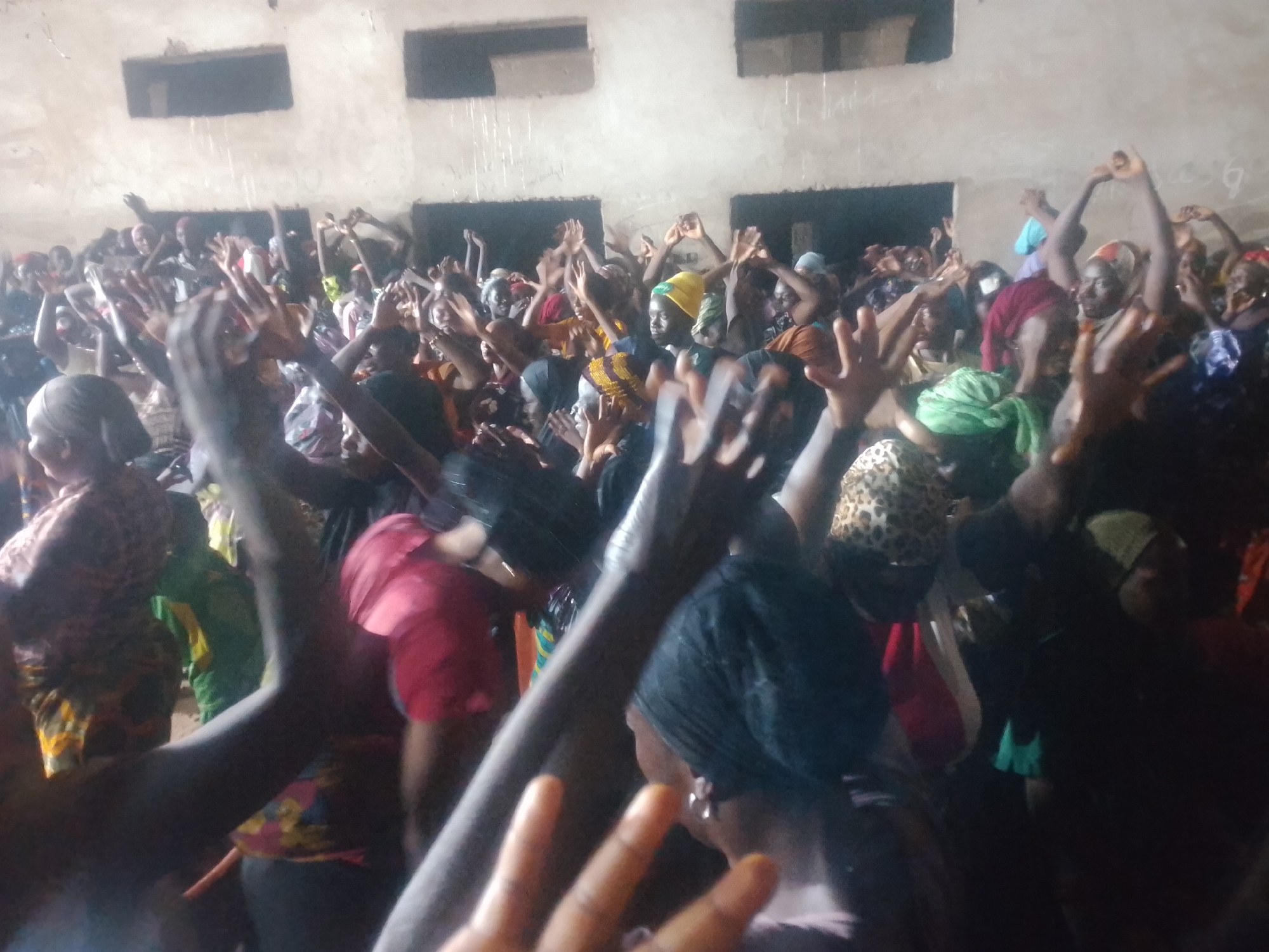 An indoor crowd of people are raising their hands in the air, some with their palms facing the camera. The lighting is dim and the scene is crowded, making it difficult to discern individual faces or actions clearly. The individuals appear to be of various ages and genders, and many are wearing head coverings and colorful clothing. The background shows a concrete wall with two rectangular openings, possibly windows or doorways. The overall impression is of a lively gathering or event. The location is described as Unknown, Unknown.