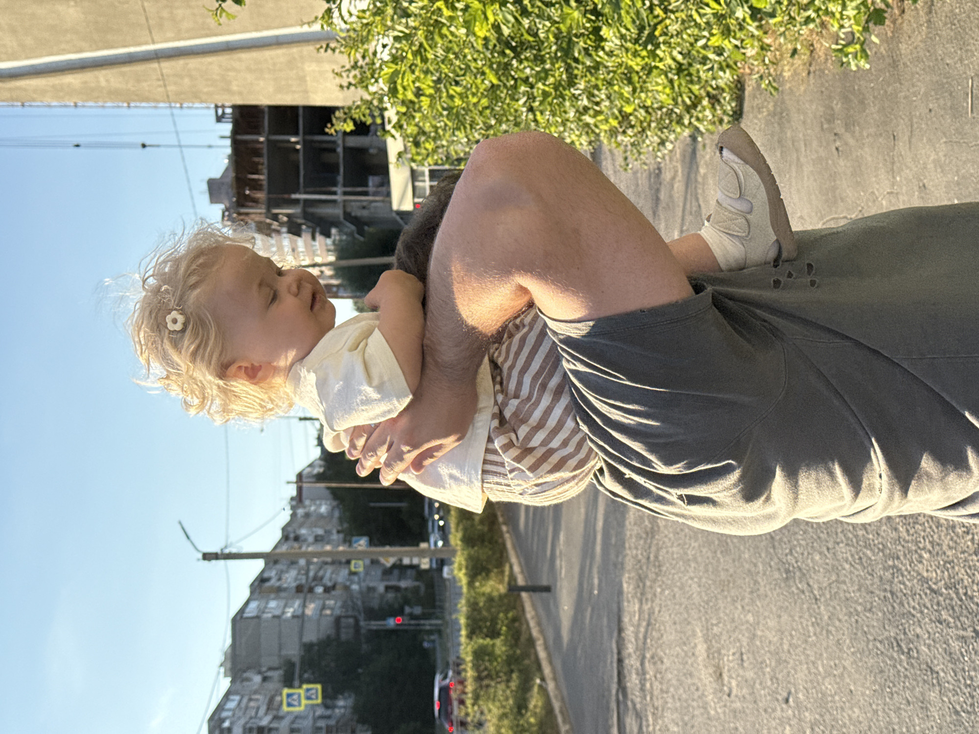 The image shows a close-up of a person holding a toddler outdoors, with a cityscape in the background. The toddler, with blond curly hair adorned with a white flower clip, is looking upwards, seemingly enjoying the moment. The toddler is wearing a light-colored top and striped shorts. The person holding the toddler has their legs bent, and is wearing a dark t-shirt and shorts. The toddler's foot, wearing a light-colored sandal, is visible resting on the person's leg.
The scene appears to be a sunny day, suggested by the bright light and shadows. The background features buildings, power lines, and some greenery, indicating an urban environment. The buildings are multi-story, characteristic of residential areas. The presence of traffic lights and what appear to be street signs suggest a street or sidewalk setting. The overall mood seems peaceful and affectionate, with the focus on the interaction between the adult and the child.
The photo is oriented vertically, with the sky at the bottom and the ground at the top, suggesting it was taken with the camera held in portrait mode, and then rotated. No visible text is present in the image.