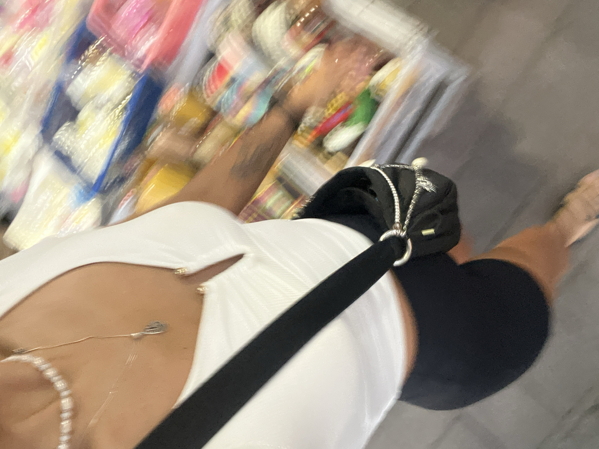 A high-angle, blurred image shows a person wearing a white top, black shorts, and a black crossbody bag with silver embellishments walking past a blurred display of packaged goods. The person is also wearing jewelry, including a pearl necklace and a delicate pendant. Part of their arm is visible, displaying a tattoo. The setting appears to be a commercial area, possibly a shop in Kuta Selatan, Indonesia, indicated by the packaged goods, but the location is not explicitly verifiable from this image. The focus is on the person and their attire, with the background details blurred and indistinct.