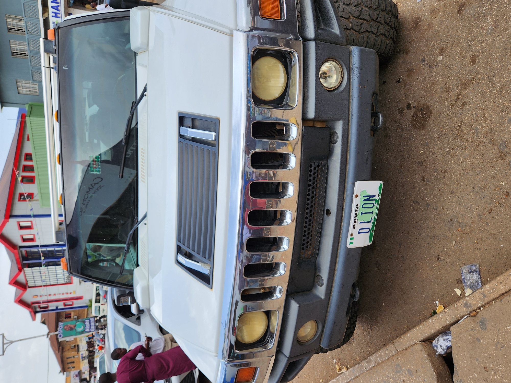 The image shows the front of a white Hummer H2 parked on a street. The vehicle has a Nigerian license plate that reads "DLION" and "ABUJA". The background includes a multi-story building with red and white accents, people standing on the sidewalk, and other vehicles parked nearby. Trash is visible along the curb.
