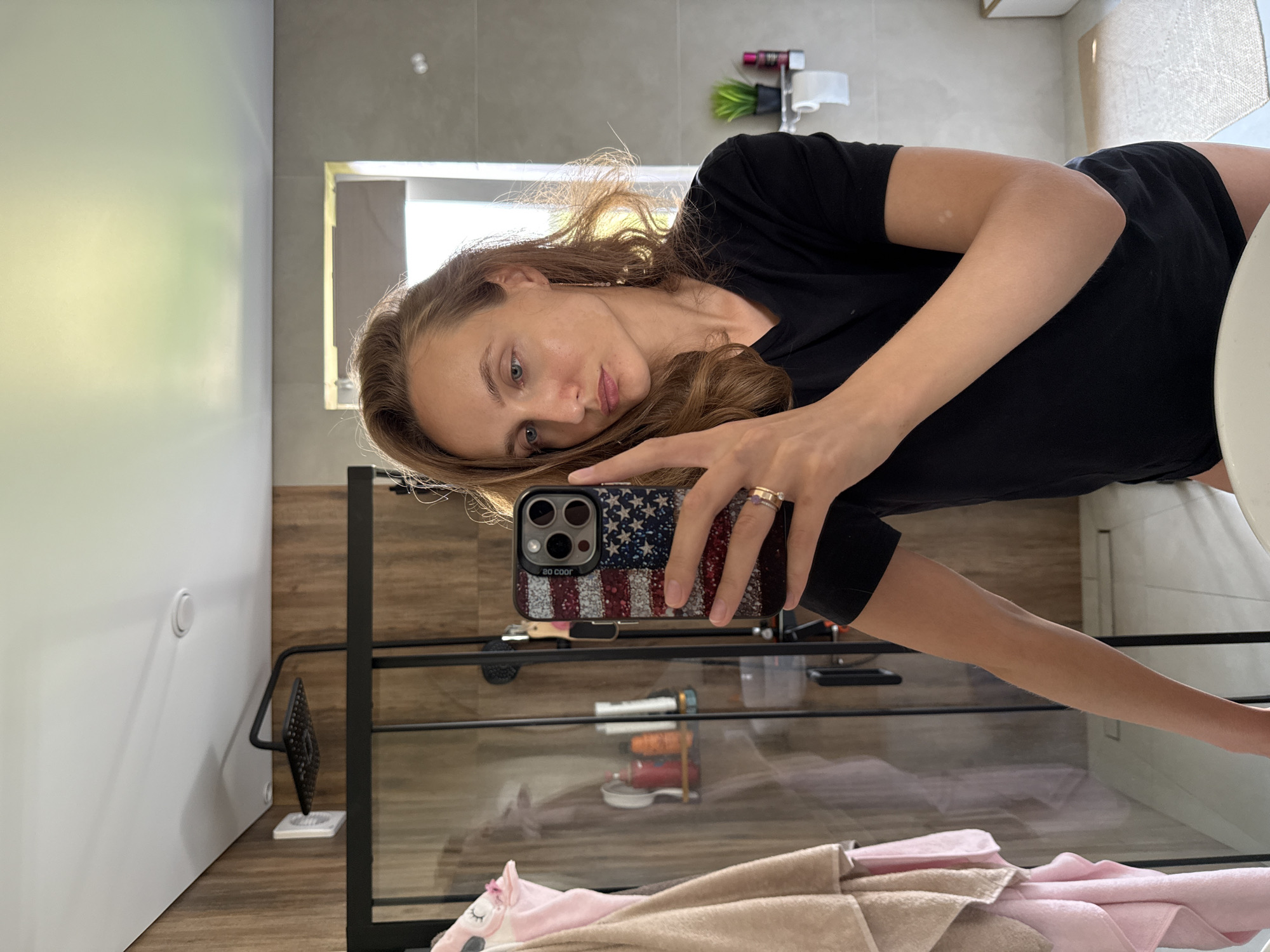 A young Caucasian woman with long, wavy brown hair and fair skin is taking a selfie in a modern bathroom. She is wearing a black t-shirt and dark shorts. She has on a ring on her left hand and a long earring in her right ear. Her phone, with an American flag-themed case, is held up to take the photo. The bathroom features wood-paneled walls and grey tiled surfaces. A shower stall with a black frame is visible to the left, with towels and toiletries inside. A small plant and a toilet paper dispenser are mounted on the wall to the right. The overall impression is one of a clean and contemporary domestic space. The location is confirmed to be in the city of Podshivalovo, Russia.