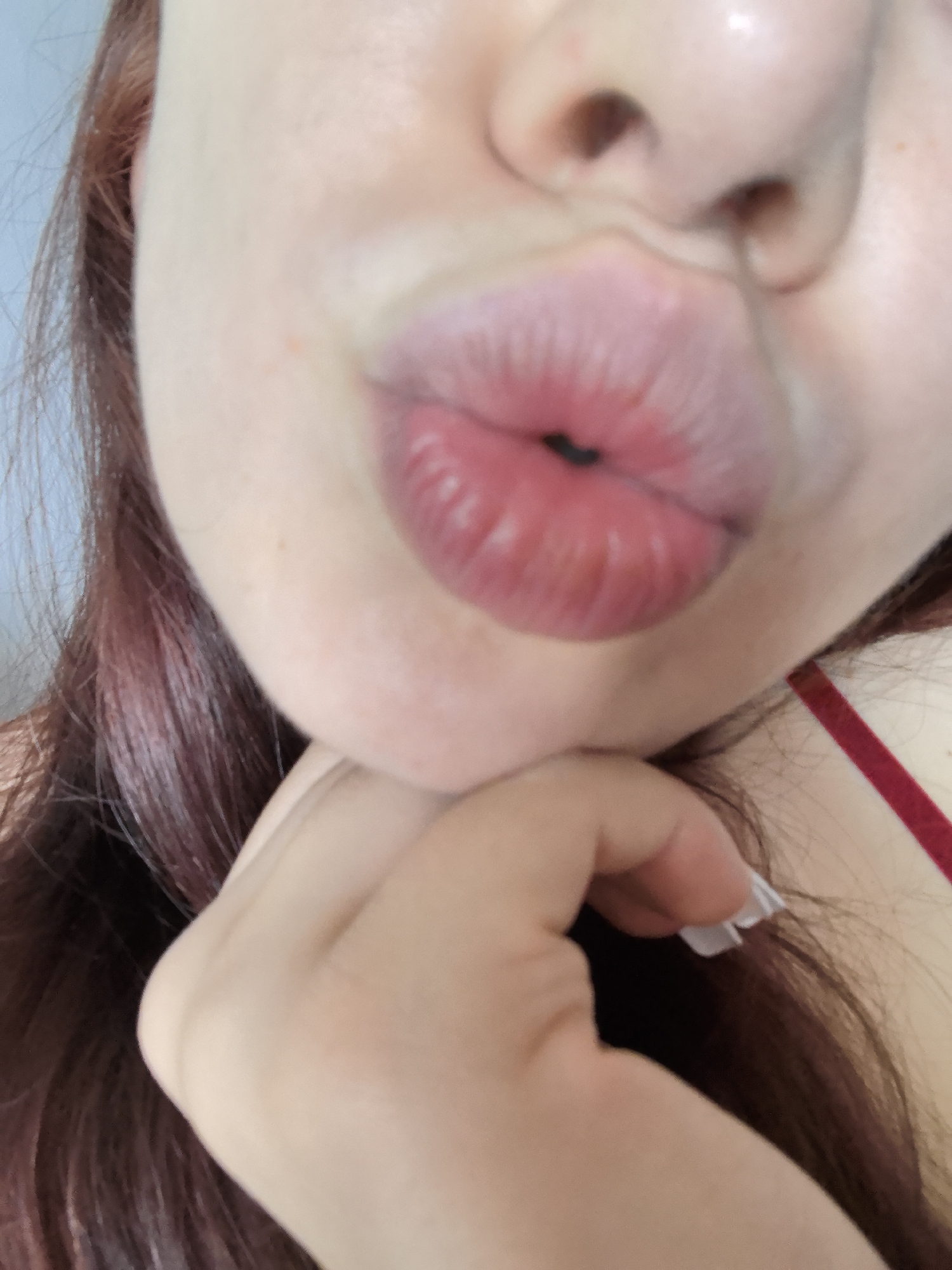 The image is a close-up, slightly blurry, portrait of a person's face, focusing on their lips pursed in a kissing gesture. The person has fair skin and reddish-brown hair that frames the lower part of their face. Their lips are plump and appear to be wearing a light-colored lip gloss or balm. The person's chin is supported by their hand, which has well-manicured nails, some of which are painted white. A thin red strap is visible on the right side of the frame, possibly part of clothing. The background is a soft, pale blue, suggesting an indoor setting. The image does not contain any elements that would allow for identification of a specific location within Stara Zagora, Bulgaria.
