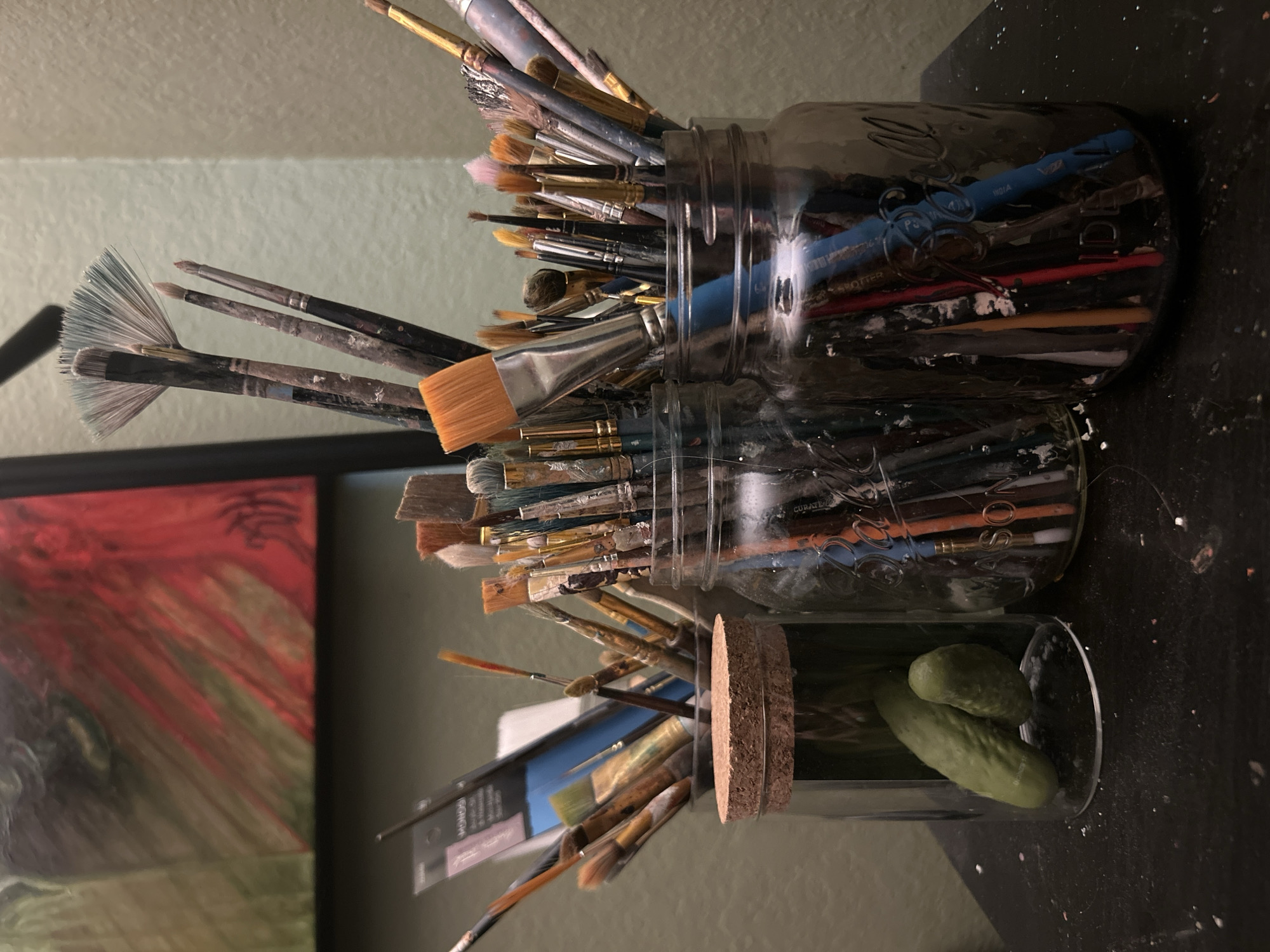 The image shows three glass jars filled with art supplies, specifically paintbrushes and drawing instruments. The jars are arranged on a dark, possibly speckled surface, likely a countertop or table. The top jar is a clear glass Mason jar packed tightly with an assortment of paintbrushes. The second jar is similar to the first, also filled with paintbrushes of varying sizes and styles. The third jar is a rectangular glass container holding a cork-like cylindrical object (possibly a jar top) and what appear to be two whole dill pickles. To the left of the jars, part of a framed painting can be seen, featuring red and green colors. The wall behind the setup is painted a pale green or gray color. The image is taken in a room with natural light. There is no indication in the image that it is in Cape Coral, Florida. There are no people visible in this image.