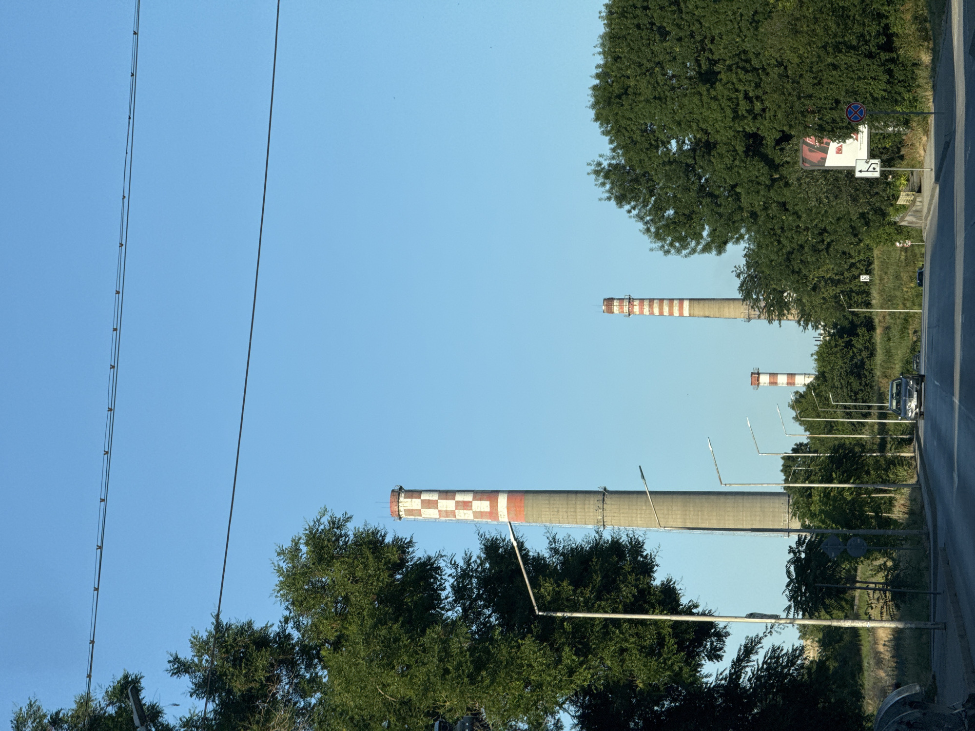 The image shows a view from a car driving on a road in Pleven, Bulgaria, with several industrial chimneys in the background. The sky is clear and blue. On the left side of the road, there are large green trees and a street lamp. Several smaller street lamps are visible along the road. In the distance, four industrial chimneys of varying heights can be seen. The closest and largest chimney is gray with red and white checkered patterns near the top. Another chimney to its right is also gray with red and white horizontal stripes. Further in the distance, two more chimneys are visible, one of which also has red and white horizontal stripes. A silver sedan is driving on the road towards the viewer. There are also road signs visible. The environment appears to be a mix of industrial and natural elements, with greenery surrounding the road and the industrial structures.