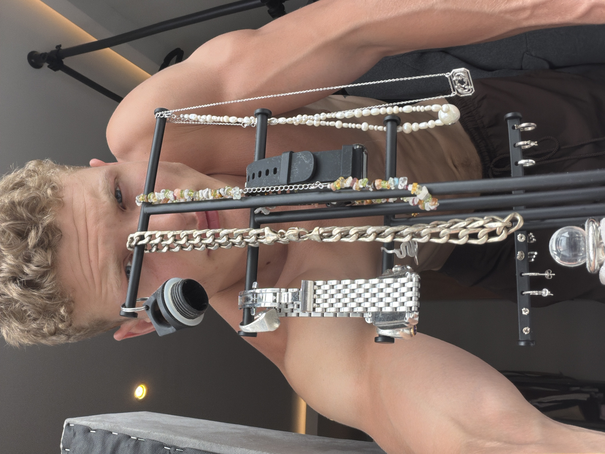 The image shows a shirtless man with curly blonde hair, viewed from a low angle and rotated ninety degrees. He is positioned behind a jewelry display stand. The display stand is black and contains several pieces of jewelry, including pearl necklaces, silver chains, beaded bracelets, a black watch band, a silver watch, and earrings. The background shows a light-colored wall and the ceiling fixture. A piece of furniture is visible in the lower portion of the image. No information confirms the location is London, United Kingdom.