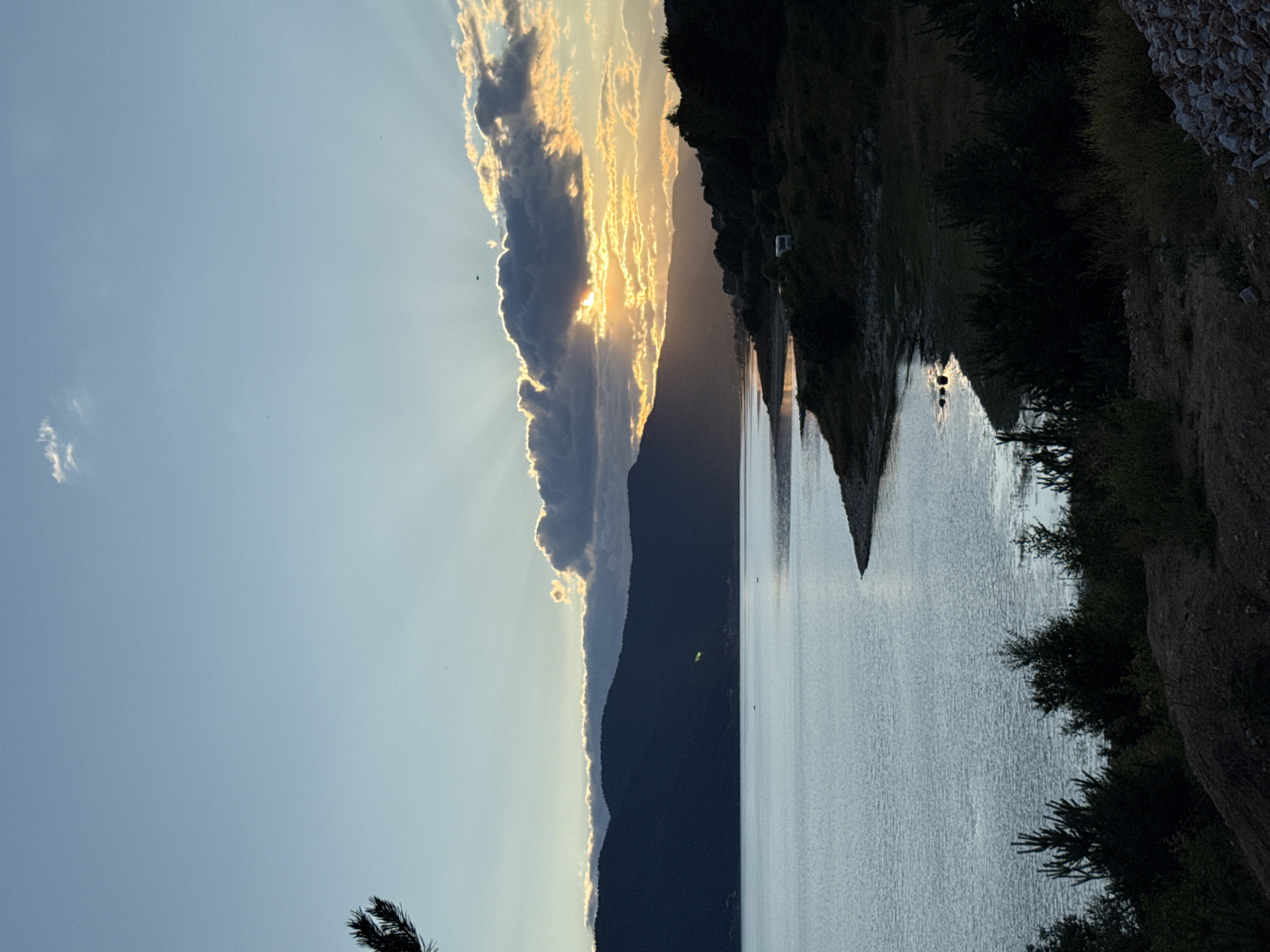 The image depicts a serene sunset over the Dospat Dam, near the town of Sarnitsa, Bulgaria. The sky is a gradient of blue, transitioning to hues of orange and yellow where the sun is setting behind a band of dark clouds. Rays of sunlight pierce through the clouds, illuminating the sky with a warm glow. In the foreground, the calm surface of the dam reflects the sky's colors. The shoreline is lined with dark green coniferous trees and shrubs, with a small embankment of rocks visible on the right. Further in the distance, a dark, undulating mountain range forms the horizon, silhouetted against the vibrant sunset. A white camper van is parked on the grassy slope near the water's edge. The overall scene is one of natural beauty and tranquility.
