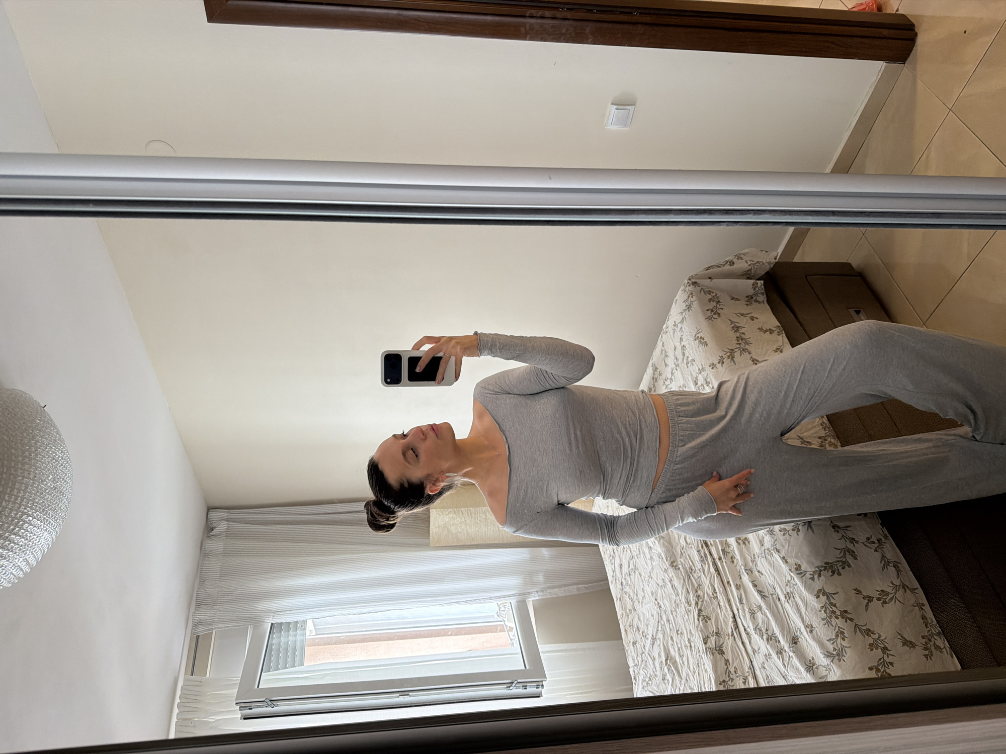 A woman is captured in a full-length mirror selfie, standing upright and holding a white smartphone in her right hand to take the photo. She has dark hair styled in a neat bun and is wearing a coordinated light grey two-piece loungewear set, featuring a long-sleeved crop top with an off-shoulder neckline and matching wide-leg trousers. A subtle necklace and a ring on her left hand are visible.
The reflection reveals a brightly lit bedroom, suggesting it is daytime. To the woman's right, a bed is covered with a white duvet adorned with a dark floral or leafy pattern, and a dark brown bed frame or headboard is visible. To her left, a window with white horizontal blinds allows natural light into the room. A portion of a modern, spherical white light fixture hangs on the wall to the far left. The walls are painted a light, neutral color, and the floor is covered in light brown square tiles. A dark wooden frame or trim runs across the top of the reflected space, and a white light switch is mounted on the wall above the bed area.
The scene depicts a personal moment of the woman taking a picture in what appears to be a comfortable and contemporary bedroom setting in Budva, Montenegro. No visible text is present in the image.