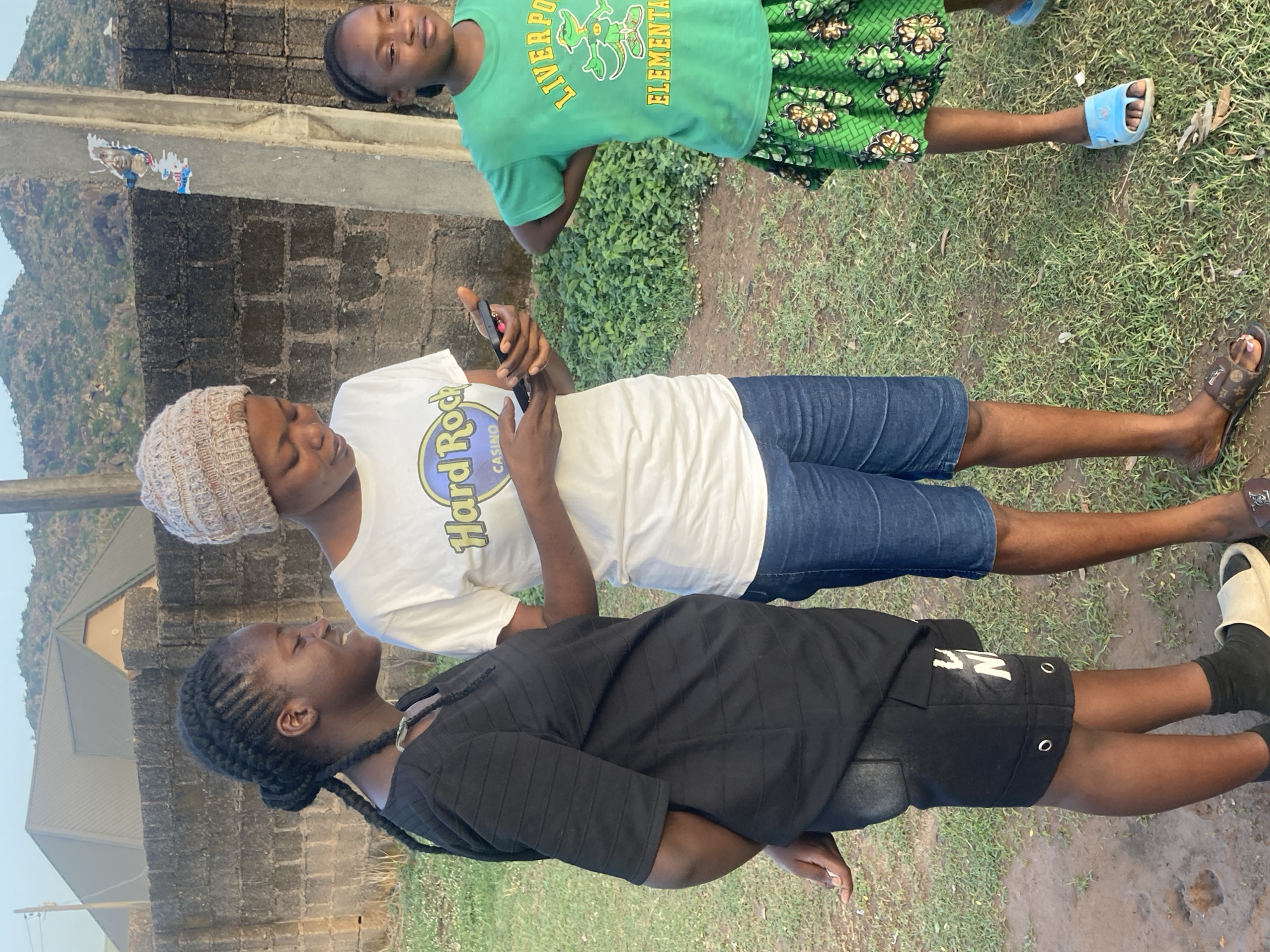 The image depicts three young individuals outdoors. One person is wearing a black top and black shorts, while another is wearing a white Hard Rock Cafe t-shirt and blue shorts. The third person is wearing a green t-shirt with the word "Liverpool" on it and a patterned green skirt. The individual in the white t-shirt is holding a phone.
The environment includes a building with brick-like patterns and green vegetation. In the background, a mountain range can be seen. Based on the context and visual cues, the location is potentially within the city of Jos, Nigeria.