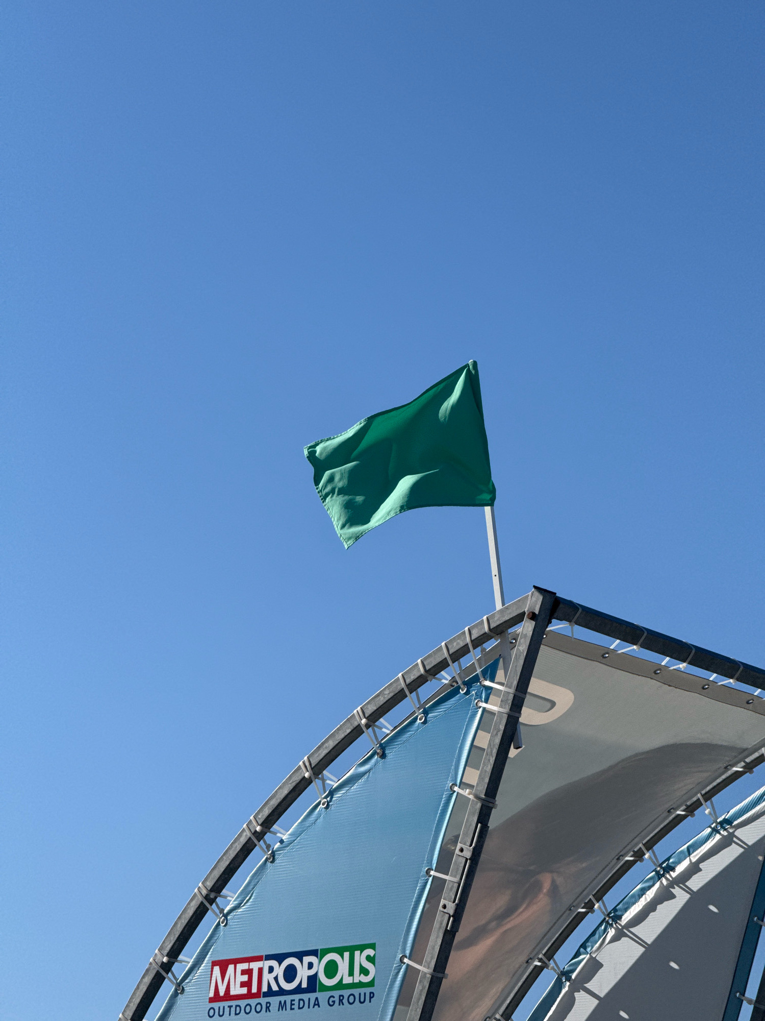 A bright green flag waves in the clear blue sky from a white flagpole. The flagpole is mounted on a metal structure that supports a large, light blue fabric banner displaying the logo "METROPOLIS OUTDOOR MEDIA GROUP" in red, white, and green lettering. The banner is attached to the metal structure with numerous white zip ties. The scene appears to be outdoors, possibly at a public venue or event.