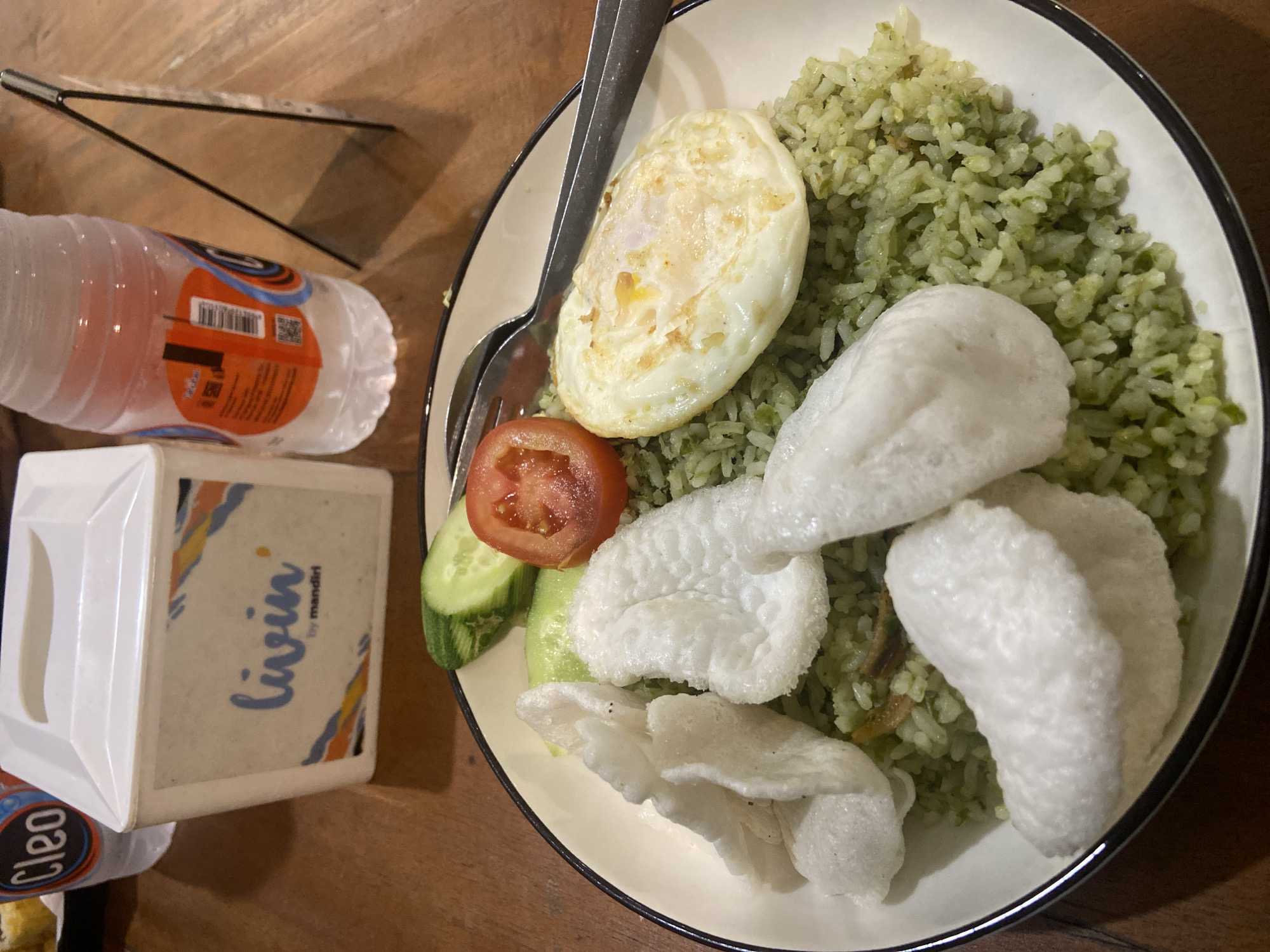 A high-angle medium shot captures a plate of food, likely nasi goreng, and a bottle of water on a wooden table. The nasi goreng is a vibrant green, topped with white crackers, a fried egg, a slice of tomato, and cucumber slices. To the left of the plate sits a white paper box with "livin by mandiri" printed on it. Behind the box, a partially visible clear bottle of water with an orange and blue label is present. The background shows a rustic wooden surface. No individuals are visible in the frame. The scene is indoors, with the lighting suggesting an ambient light source, possibly from above.