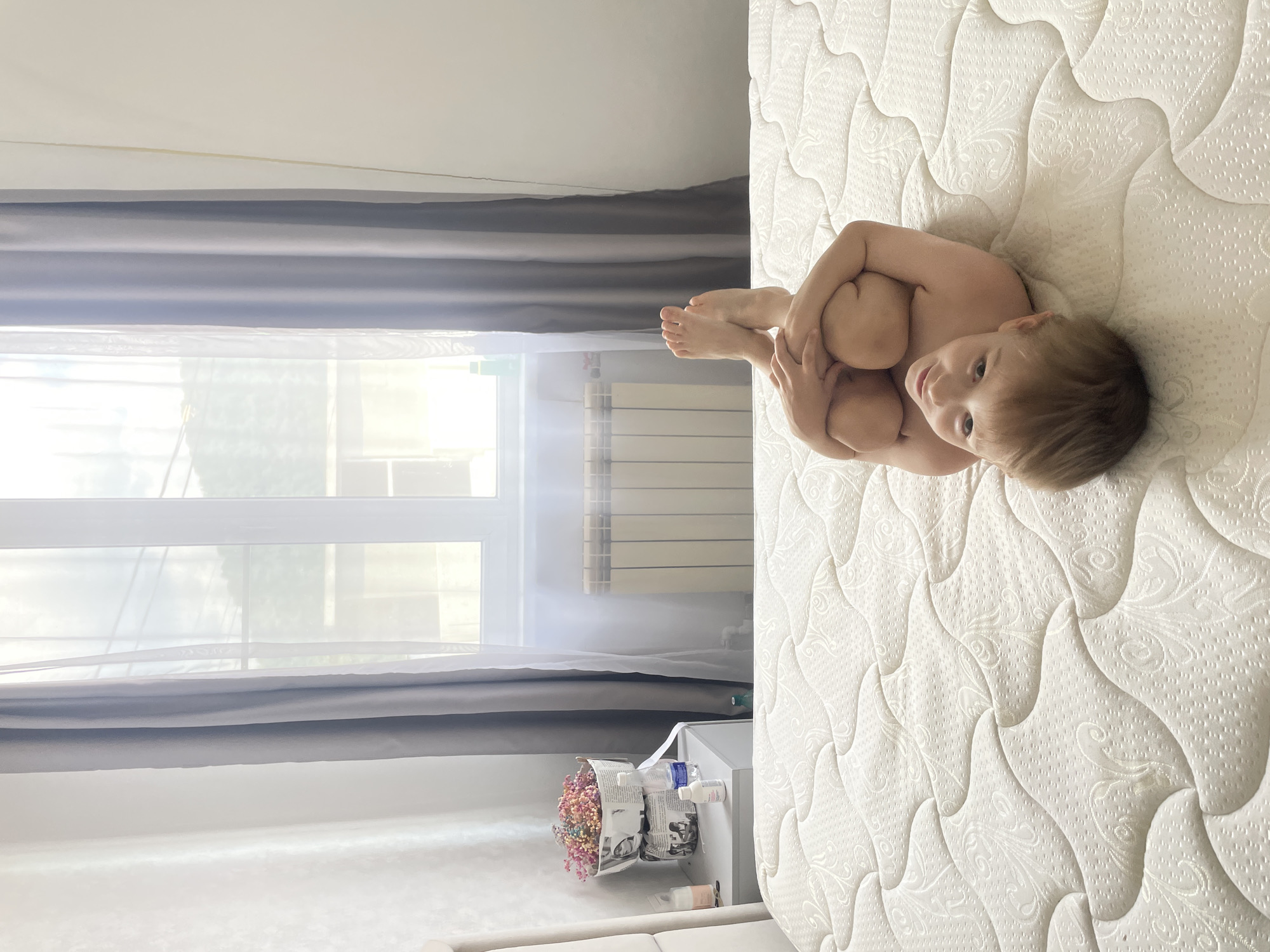 A brightly lit indoor scene depicts a young child lying on a white quilted mattress in a bedroom, identified as being in Baranovka, Russia. The child, a toddler with light blonde hair and light eyes, is positioned centrally on their back, with knees bent and arms wrapped around their legs, looking directly at the camera with a neutral expression. The child appears to be unclothed.
The mattress has an intricate pattern of raised swirls and dotted textures. To the left of the frame, a segment of a beige upholstered headboard is visible, next to a light grey nightstand. On the nightstand, two stacked decorative containers, seemingly crafted from newspaper with printed text and images, hold an arrangement of colorful dried or artificial flowers in shades of pink, orange, blue, and white. Beside these, two plastic bottles (one larger clear bottle with a blue cap, and one smaller white bottle with a blue cap) and a small white jar are placed.
The background features a large window with white frames, covered by sheer white curtains and thicker grey blackout curtains. Below the window, a white horizontal panel radiator is mounted against a light-colored wall. The room is illuminated by strong natural light entering through the window, creating some overexposed areas in the upper background.