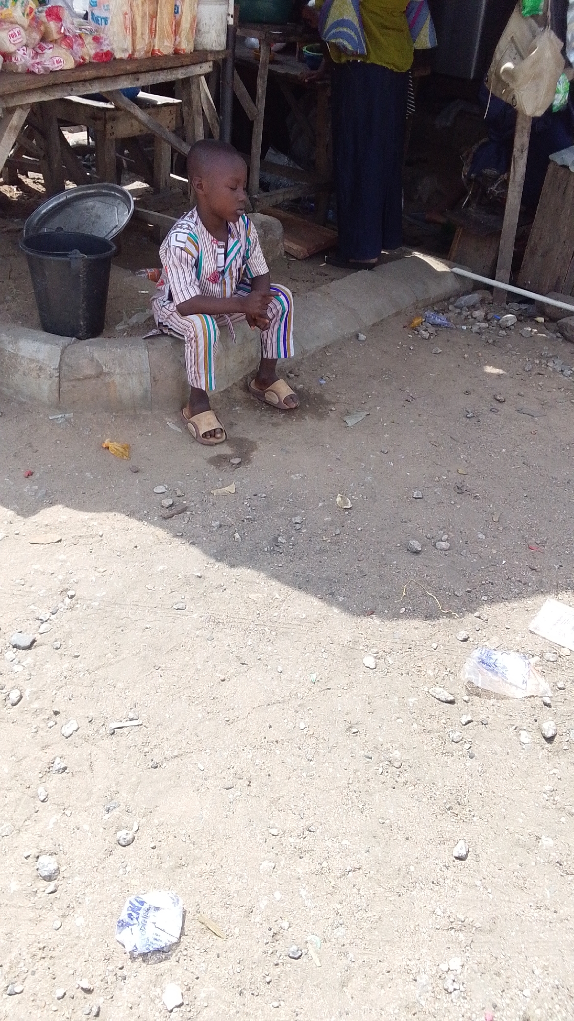 The image shows a young boy sitting on a curb in Lokoja, Nigeria. He is wearing a striped, multicolored outfit and sandals. A black bucket sits next to him on the curb. Behind the boy is a makeshift wooden structure with goods on display, likely a market stall. An adult partially visible behind the stall is wearing a yellow top and dark skirt. The ground is dirt and scattered with debris.