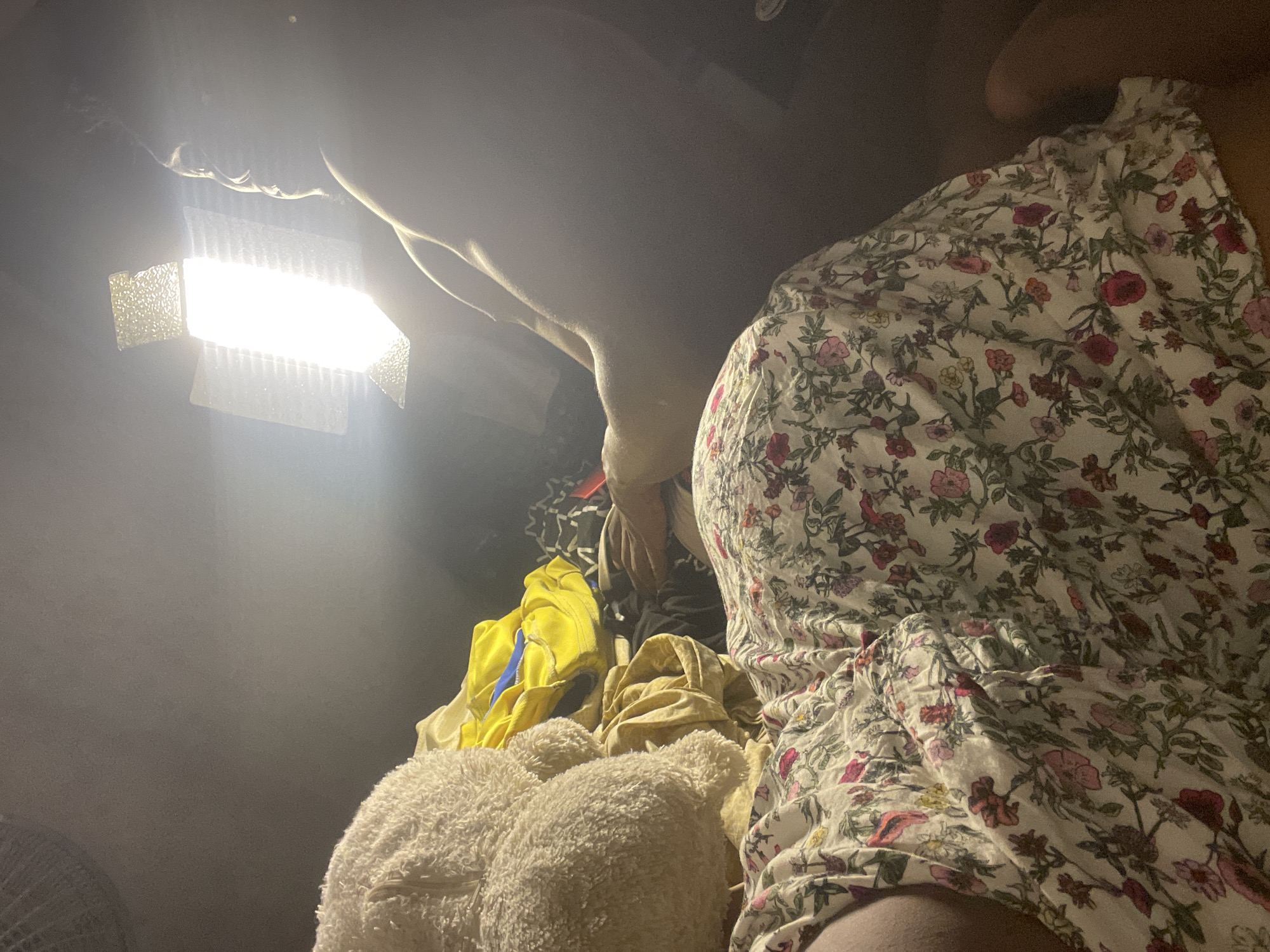 The image shows a person wearing a floral print dress sitting next to a pile of clothes and a teddy bear. The clothes are a mix of colors, including yellow and beige. A bright light source is positioned above the person. The location is unspecified, and there is no indication this image was taken in Lokoja, Nigeria.