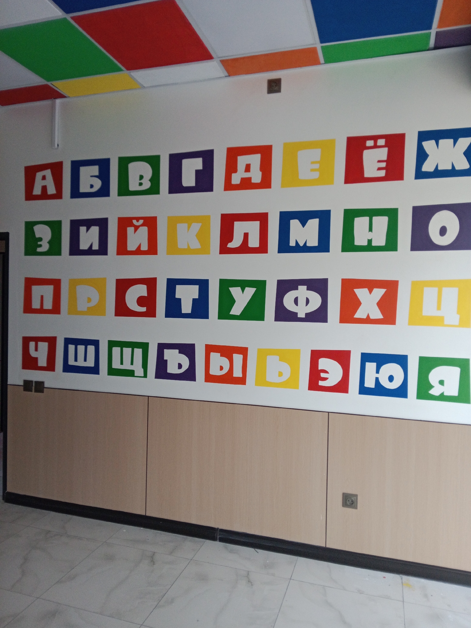The image shows a wall decorated with the Cyrillic alphabet, with each letter inside a colored square. The letters are arranged in rows. Above the alphabet wall, a colorful ceiling with geometric patterns is visible. Below the alphabet, a light-colored wooden cabinet with black trim runs along the wall. The floor is tiled with large, light-colored tiles. An electrical outlet is visible on the cabinet. This scene appears to be in a classroom or a children's learning space. Without further context or identifying markers, the specific location in Tashkent, Uzbekistan cannot be determined.