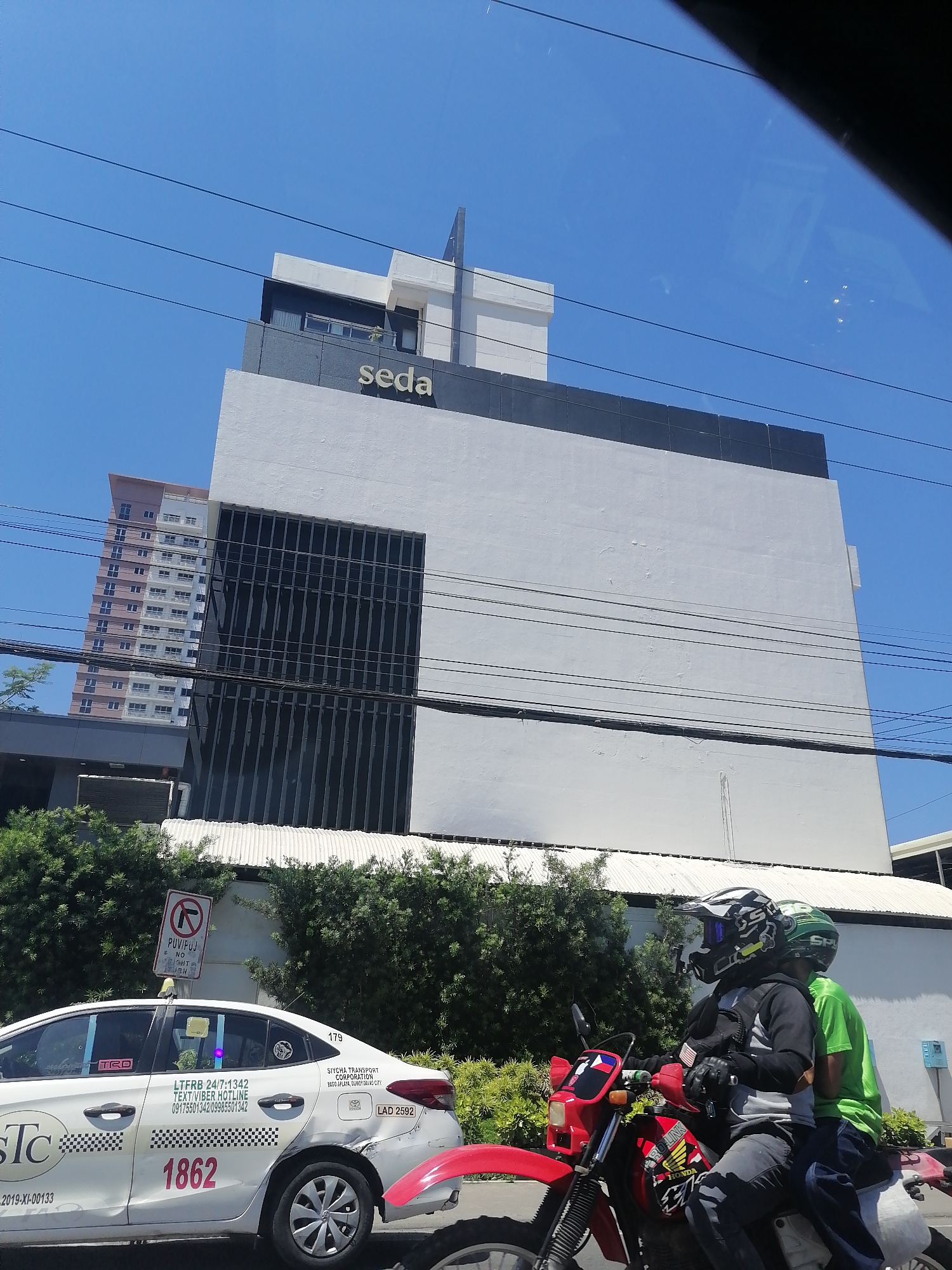 In Davao City, Philippines, a bright sunny day is depicted, with a clear blue sky dominating the upper portion of the frame. A multi-story building, identified by the prominent "seda" signage, stands as the central subject. This building features a modern architectural design with a predominantly white facade, accented by dark horizontal bands and a section of vertical dark windows. To the left of this building, a taller, pink and white residential building is partially visible.
In the foreground, a white sedan taxi, marked with the number 1862 and the "STC" logo, is parked. The taxi has various decals and license plates, including "LTO" and "LAD 2592". A red motorcycle with two riders, both wearing helmets, is positioned next to the taxi, also facing forward. The rider in front is wearing a black and white helmet and a grey hoodie, while the passenger behind wears a green helmet and a green shirt. The motorcycle has various stickers and a Philippine flag attached to the handlebars.
The environment surrounding the vehicles and buildings includes lush green trees and hedges, suggesting an urban or suburban setting. Overhead, several black electrical wires crisscross the sky, indicating an established infrastructure. A traffic sign with a red circle and a diagonal bar, prohibiting a left turn, is mounted on a pole near the taxi. The overall scene captures a typical day with vehicles and buildings under a clear sky.