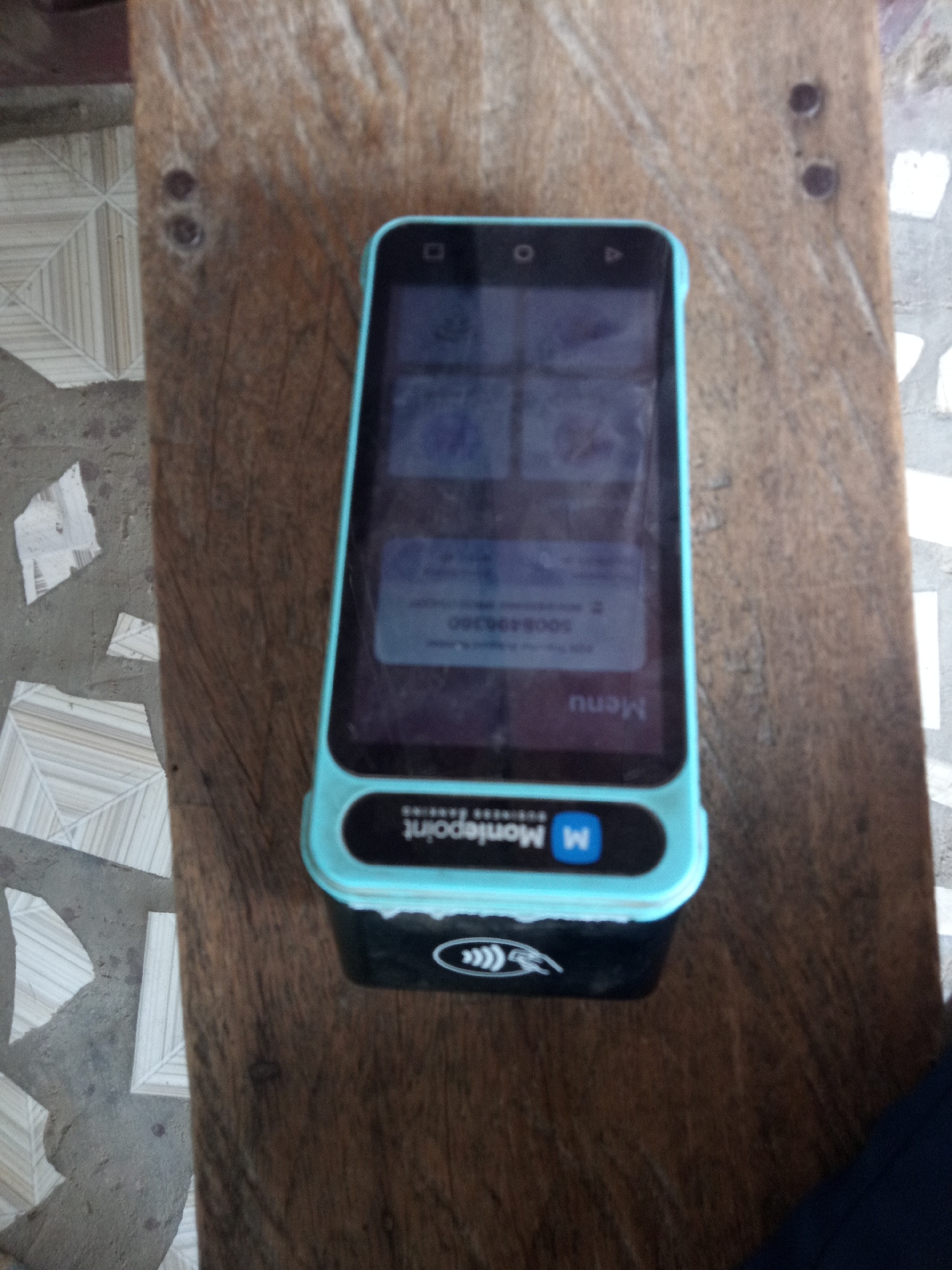 The image shows a close-up, top-down view of a blue and black mobile point-of-sale device resting on a textured wooden surface. The device has a touchscreen display that shows a dimly lit screen with several abstract images or icons. Below the screen, a button labeled "Menu" is visible. The bottom of the device features a black section with a white logo that includes the letter "M" inside a blue square and the word "Moniepoint" written in white letters. There are also some wavy lines above the "Moniepoint" text, possibly indicating a contactless payment symbol. The wooden surface appears weathered and has two dark circular indentations, suggesting it might be a table or countertop. The background is blurred, but hints of patterned flooring and white surfaces can be seen. There is no visible indication of the time of day, weather, or specific emotions.