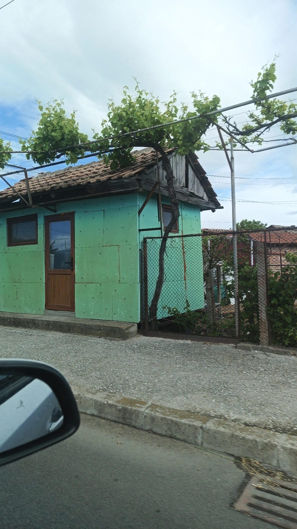 The image shows a small, green building in Milevo, Bulgaria. The building is constructed with green fiber cement panels and has a brown tiled roof. A wooden door is visible on the front, along with two small windows. A metal frame supports a vine with green foliage above the building. To the side of the building, there is a chain-link fence, with some greenery behind it. Part of a car’s side mirror is visible in the lower-left corner. The building sits on a concrete foundation and is adjacent to a pebbled area and a curb, with a street visible. The sky is partly cloudy.