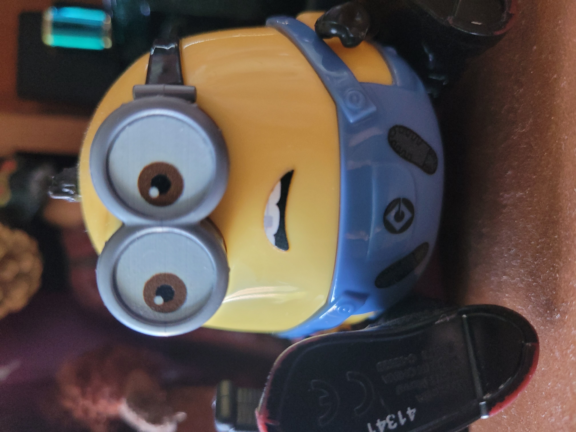 A close-up, eye-level shot shows a yellow Minion toy with large, round goggles and a blue jumpsuit. The Minion's mouth is open in an expression of surprise or distress. Part of a black shoe with red trim and some lettering is visible in the lower left corner. The background is blurred and appears to be indoors with dark colors and some out-of-focus objects. The scene is not located in Khimki, Russia.