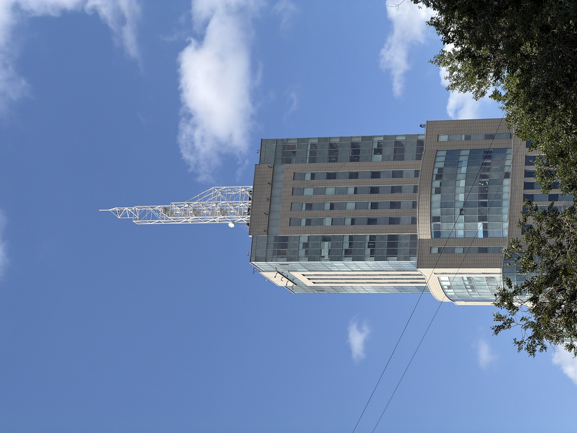 A close-up, low-angle exterior shot captures the upper portion of a modern skyscraper in Moscow, Russia, against a bright blue sky with scattered white clouds. The building features a beige facade with large, rectangular glass windows. The top of the building is crowned with a tall, white, lattice-like antenna or radio mast, reaching high into the sky. The foreground is partially obscured by the dark green foliage of trees, suggesting an urban park or street setting. The image composition focuses on the architectural elements of the building and its prominent antenna, set against a clear day.
