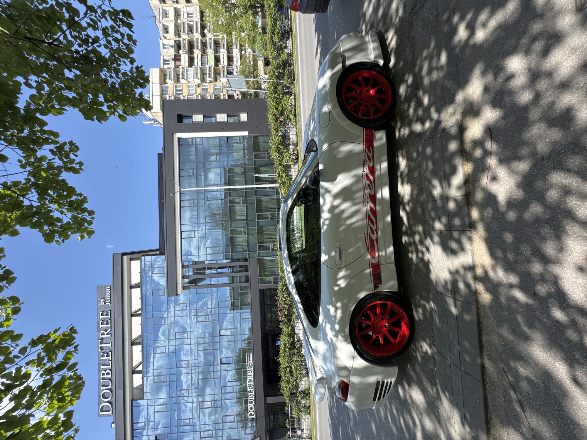 The image depicts a white Porsche Carrera parked on the street in Plovdiv, Bulgaria. The car features a red "Carrera" stripe along its side and red wheels. In the background is the DoubleTree by Hilton Plovdiv hotel, a modern glass building with the hotel name prominently displayed. Adjacent to the hotel is a taller, more conventional apartment building. The weather appears to be sunny with a clear blue sky. Shadows from nearby trees are cast on the street and the car. The image is taken at a low angle, emphasizing the height of the buildings in the background.