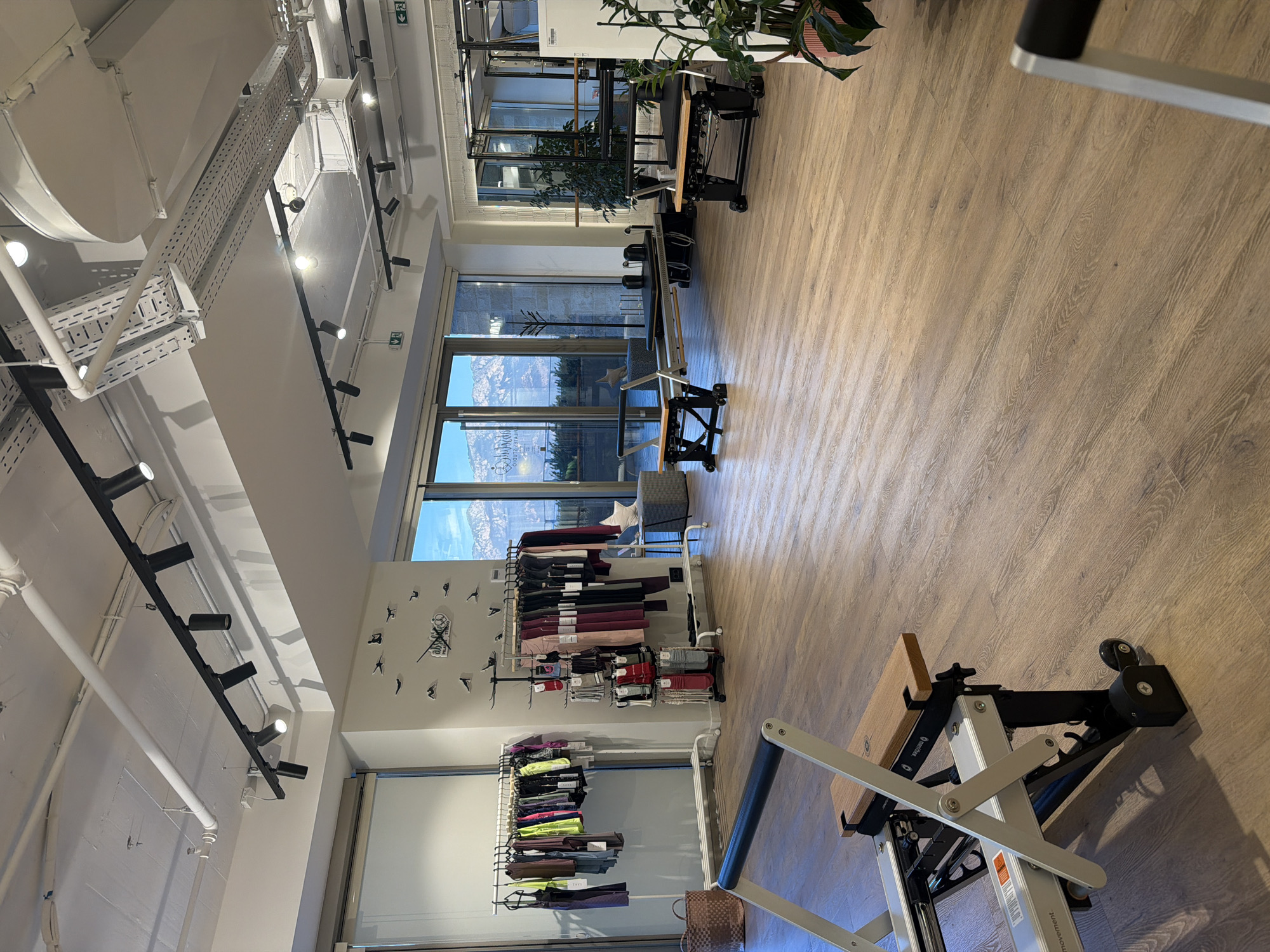 The image depicts the interior of a Pilates studio located in the city of Boreti, Montenegro. The studio features a spacious, open-plan layout with light-colored wooden flooring. Large windows offer a view of a mountainous landscape, suggesting a scenic location.
Prominent in the foreground is a piece of Pilates equipment, likely a Reformer, characterized by its white metal frame, wooden footboard, and black upholstery. Another similar apparatus is visible further into the room.
Along the walls, clothing racks are stocked with athletic wear, including leggings and tops, indicating the studio also sells apparel. A tall rack displays various pairs of socks. Decorative elements include a large clock with bird-shaped hour markers on one wall and several potted plants, adding a touch of greenery to the space. Overhead, track lighting illuminates the studio. The overall impression is one of a modern, well-equipped, and aesthetically pleasing fitness environment.