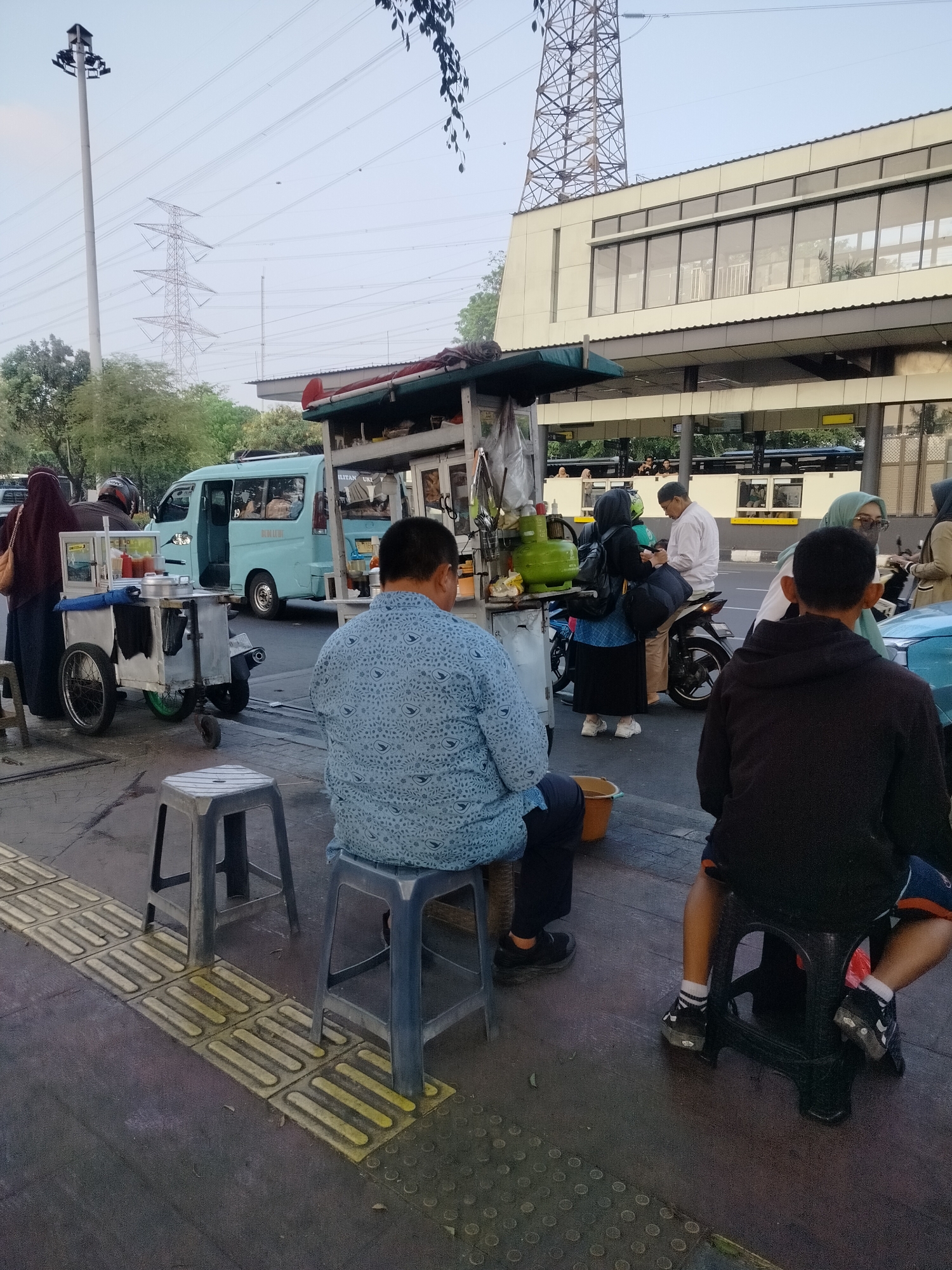 A street scene is depicted in East Jakarta, Indonesia, likely near a transit hub given the presence of a blue public transportation van and a modern building with large windows. In the foreground, several individuals are either seated or standing around street food carts. A man wearing a blue, patterned shirt is seated on a stool, facing away from the camera, with his back to a food cart that has a blue tarp. To his right, another person is seated on a black stool, also facing away, wearing a dark hoodie and shorts. A woman in a hijab is visible near the food cart, appearing to interact with it. A man in a white shirt and cap is seated on a motorcycle. Further back, a blue van with its door open is parked. The environment includes a sidewalk with tactile paving, a road with a few vehicles, and power lines and towers in the background. The lighting suggests it is either early morning or late afternoon.