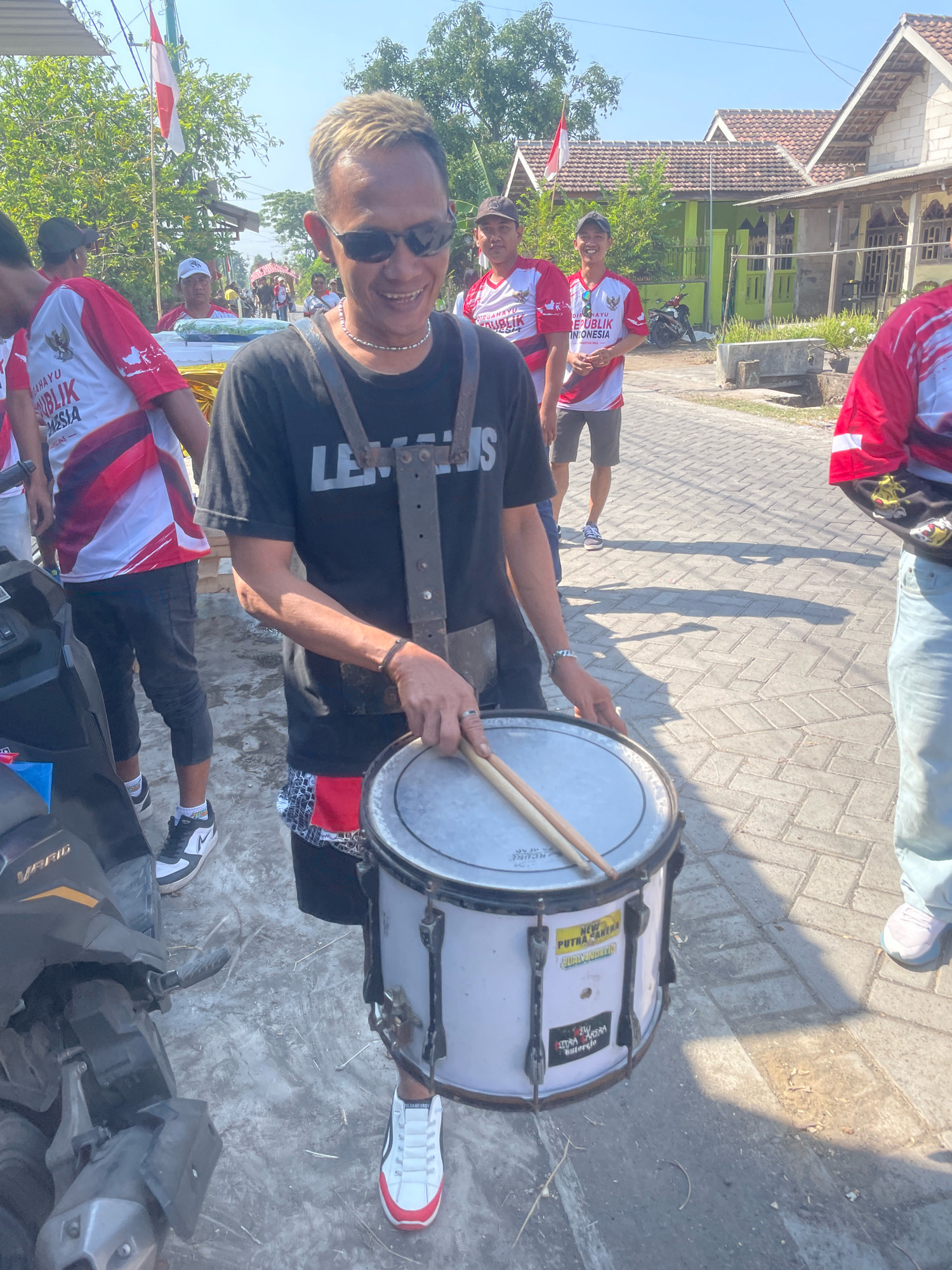 In the city of Kebon Waris, Indonesia, a man with bleached hair and sunglasses is playing a snare drum. He is wearing a black t-shirt with the word "LEGALIS" printed on it in white, and a strap system across his chest. He holds two drumsticks, one resting on the drum's rim. The drum is white with black hardware and has stickers on its side, one of which reads "NEW PUTRA KANERA SUMEDANG". He is wearing black shorts with a red and white patterned hem, and white sneakers with black accents. The man is standing on a paved street with a brick pattern. Behind him, other people are visible, some wearing red and white shirts with Indonesian flags. In the background, there are houses with tiled roofs, green vegetation, and trees. A motorcycle is parked to the left of the frame. The sky is clear and blue, suggesting a sunny day.
