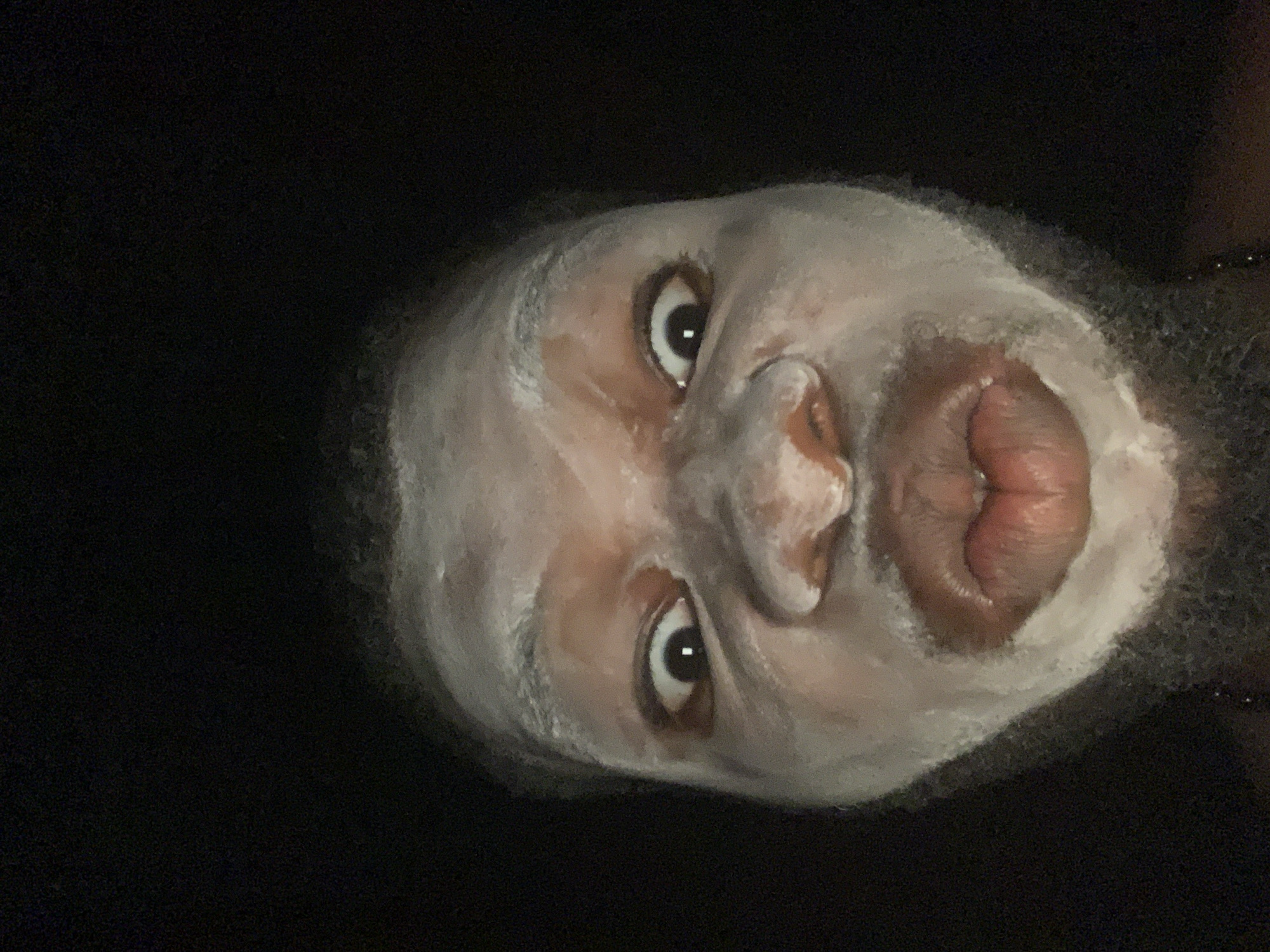 A close-up shot of a person's face with a white mask applied. The person is looking directly at the camera with wide eyes and puckered lips. The background is dark, and the face is angled. No information indicates this image was taken in Onitsha, Nigeria.