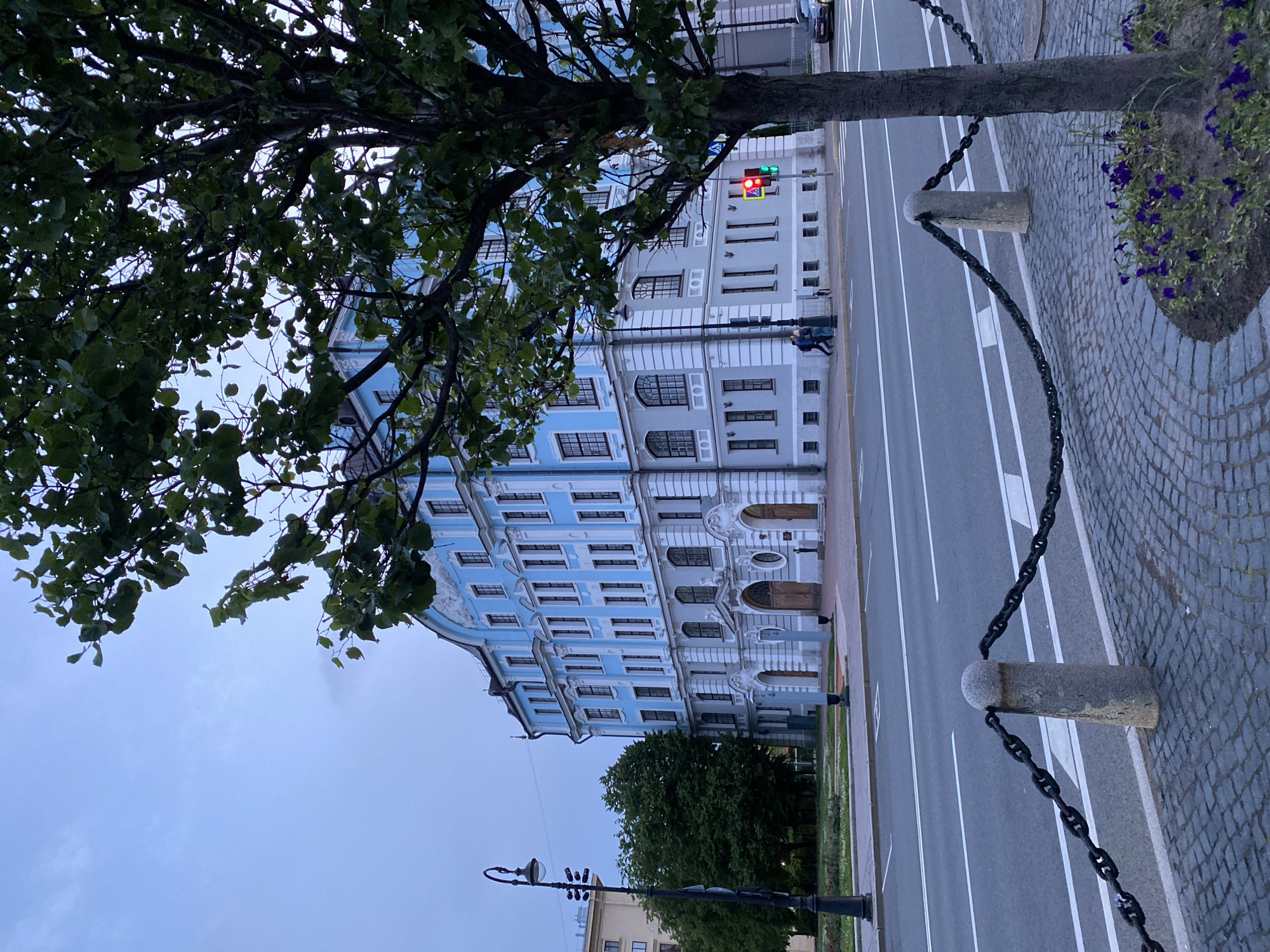 This is a wide shot of a large, light blue building in Saint Petersburg, Russia, under a cloudy sky. The building has a classical architectural style with white ornate trim, numerous windows, and a curved roofline. In the foreground, a tree with green leaves partially obscures the view of the building. A street with traffic lights, a crosswalk, and a paved sidewalk with stone bollards and chains runs in front of the building. A few people are walking on the sidewalk, and a car is visible in the distance. The overall scene suggests a late afternoon or early evening setting in an urban environment.