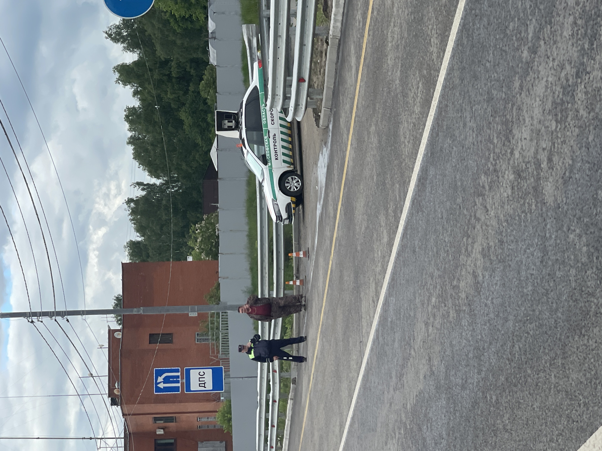 A traffic police officer is seen writing a ticket for a male civilian near the road in Taraskovo, Russia. The officer is wearing a dark blue uniform, and the male civilian is wearing a red shirt with a camouflage jacket. Beside them, a traffic police patrol car is parked on the shoulder. Traffic cones are set to provide a safety barrier. The surroundings include a roadside barrier, a gray asphalt road, and in the background, a building with a "DPS" sign on the wall and a row of green trees.