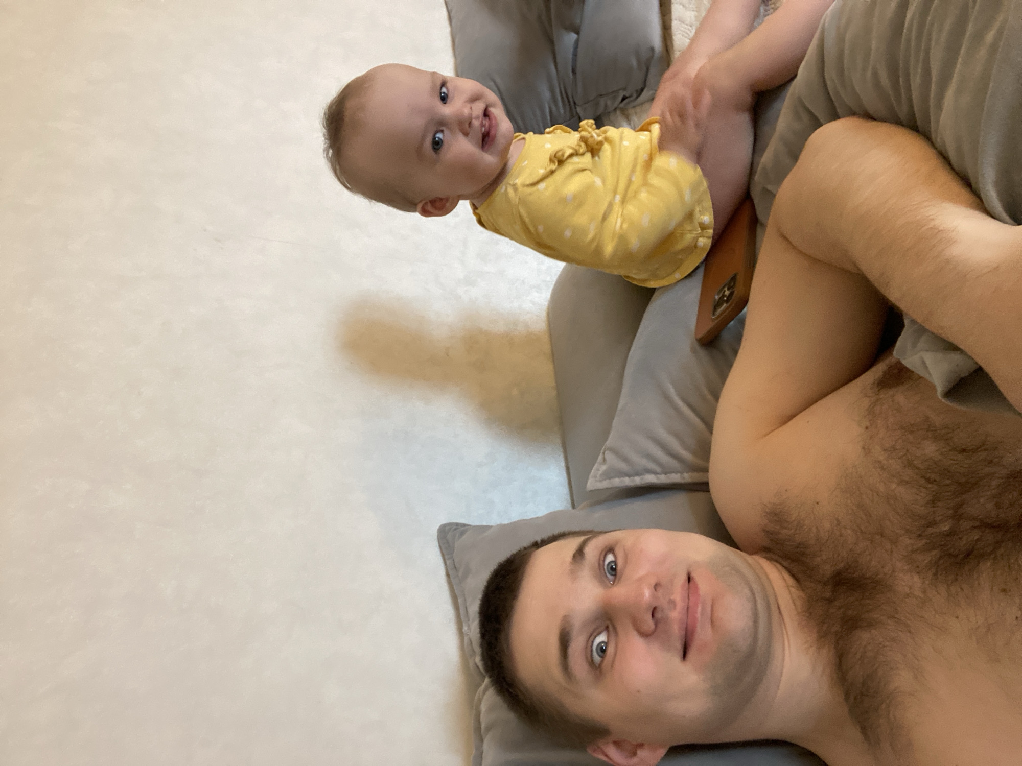 This media file captures an intimate indoor scene featuring a man and a baby/toddler, likely a father and child, in Chaikovsky, Russia. The image is taken from an overhead perspective, with both individuals lying down and looking directly at the camera, suggesting a playful moment or a selfie.
The man, positioned in the lower right, is shirtless, revealing significant chest hair. He has short, dark hair and blue eyes, and his expression is relaxed, with a slight smile. He is resting his head on a grey pillow.
Beside him, in the upper center, is a baby or young toddler. The baby is wearing a bright yellow romper or dress with white polka dots and ruffles. The baby also has blue eyes and is smiling broadly, showing a happy and engaging demeanor. The baby's head is tilted, giving a cheerful appearance. A brown smartphone is partially visible near the baby's legs, tucked into a cushion.
The background consists of a plain, light-colored surface, possibly a floor or wall, with subtle texture, and grey pillows, reinforcing the indoor setting. The lighting is soft, indicative of an indoor environment. No text is visible in the image. The overall mood is warm and affectionate.
