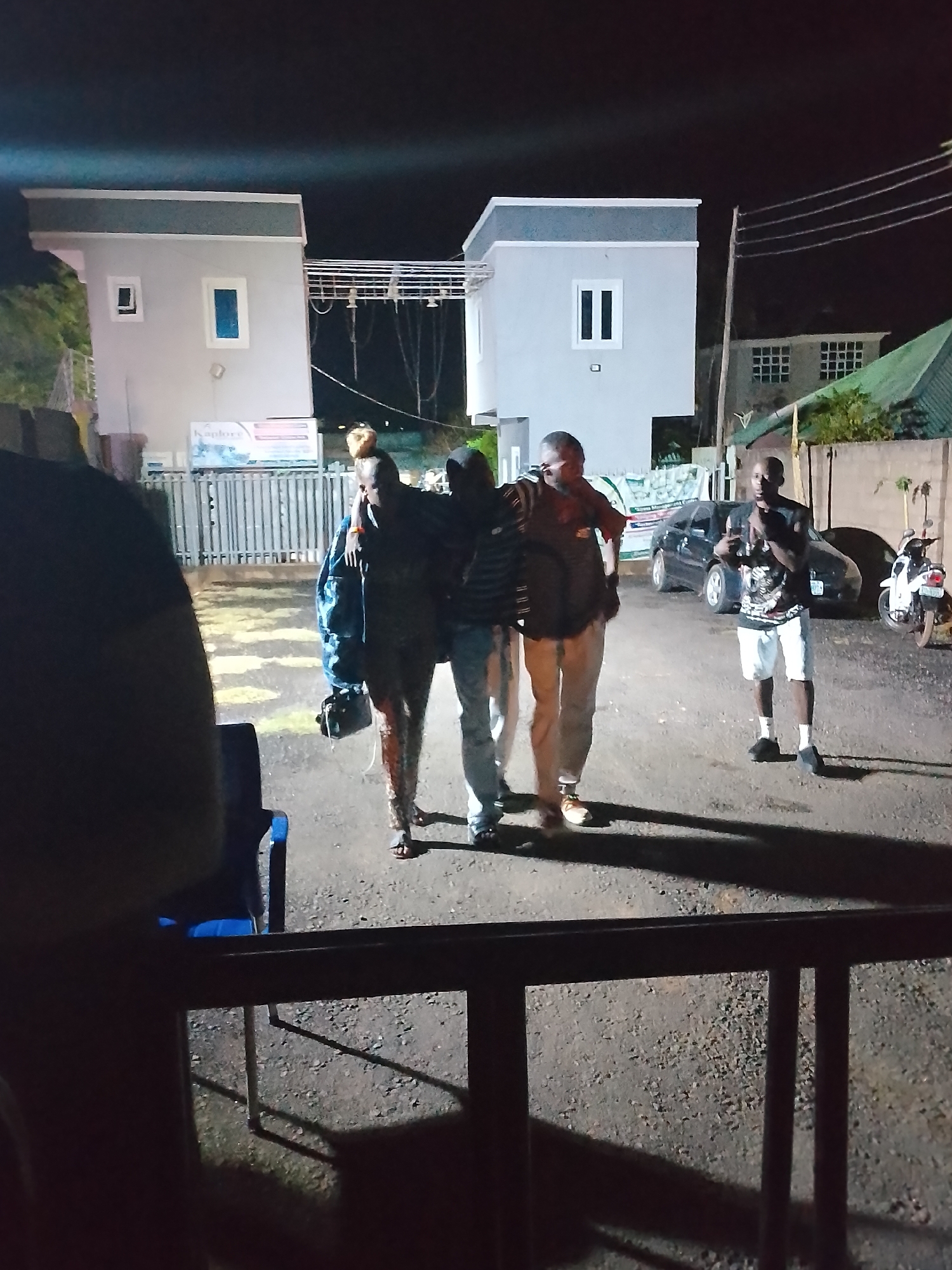 The image, taken at night in what appears to be Narayi, Nigeria, shows four individuals outdoors. Three of them are linked arm-in-arm in the center of the frame. To their right, a fourth person stands separately. Behind them are two two-story buildings with a fence between them. A car and a motorcycle are parked near the fourth person. The foreground is partially obscured by a dark railing, giving the impression the image was taken from a slightly elevated position. The ground appears to be gravel or asphalt.