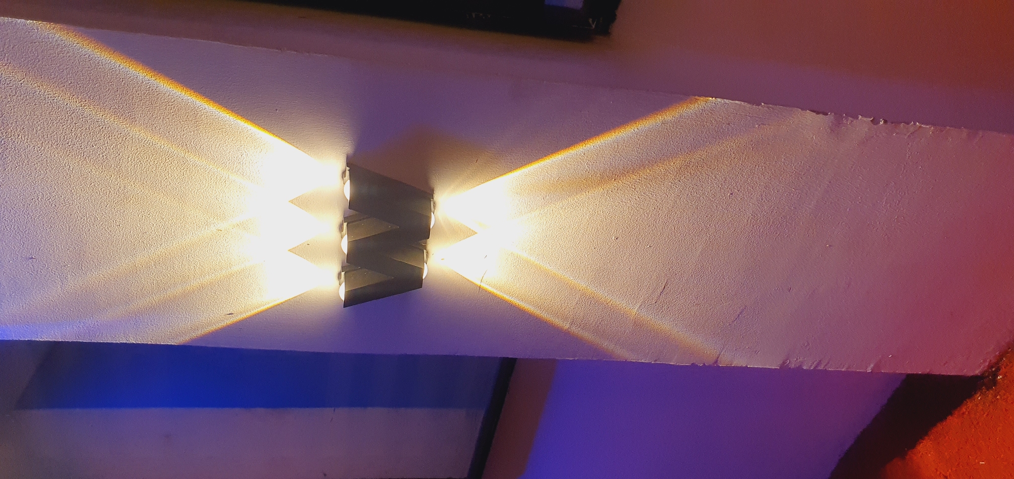 The image shows a close-up of a textured white wall with a "W" shaped light fixture mounted on it. The fixture has three bulbs that cast beams of light upwards and downwards, creating a geometric pattern of illumination on the wall. A blue light source is visible on the left side of the frame, casting a shadow on the wall. The overall lighting is dramatic, with strong contrasts between light and shadow. There is a hint of red on the floor in the bottom left corner. The image is likely taken indoors in an urban setting, possibly a modern building or a decorative installation. There are no people visible in the image. The location is Gura Top, Nigeria.