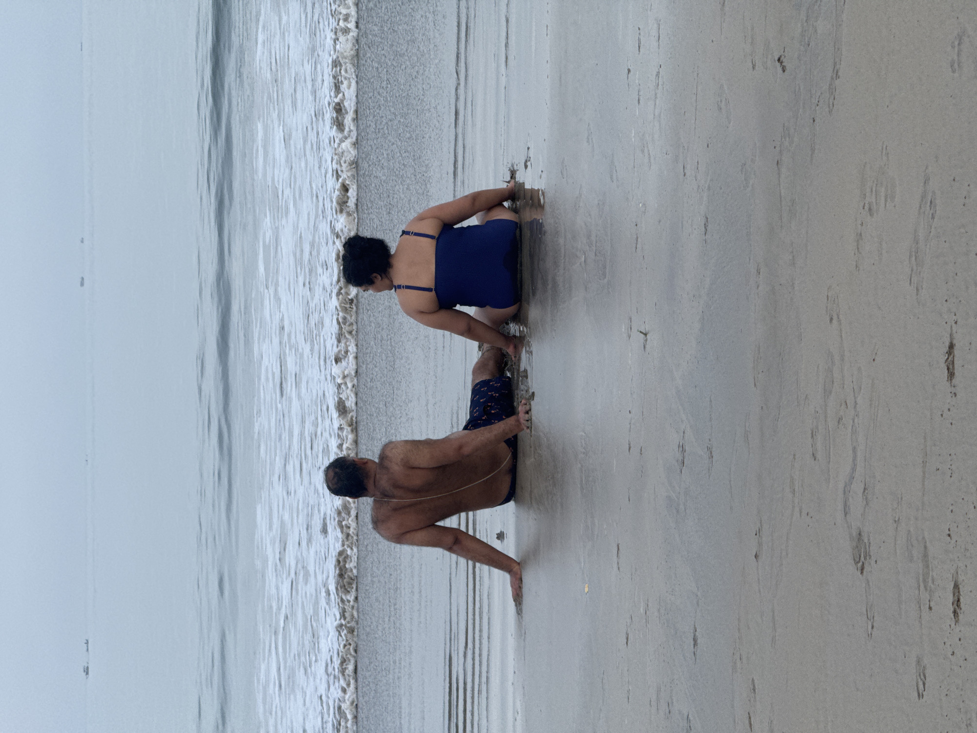 The image captures two individuals, a man and a woman, positioned near the shoreline of what appears to be a beach. The woman, clad in a blue swimsuit, is crouched down. The man, wearing dark-colored swim trunks, is positioned to the right of the woman, also crouched down. The sea is visible on the left, with waves lapping onto the sandy beach. The sky is overcast. The location may be Jimbaran, Indonesia, based on visual characteristics of the beach and sea.