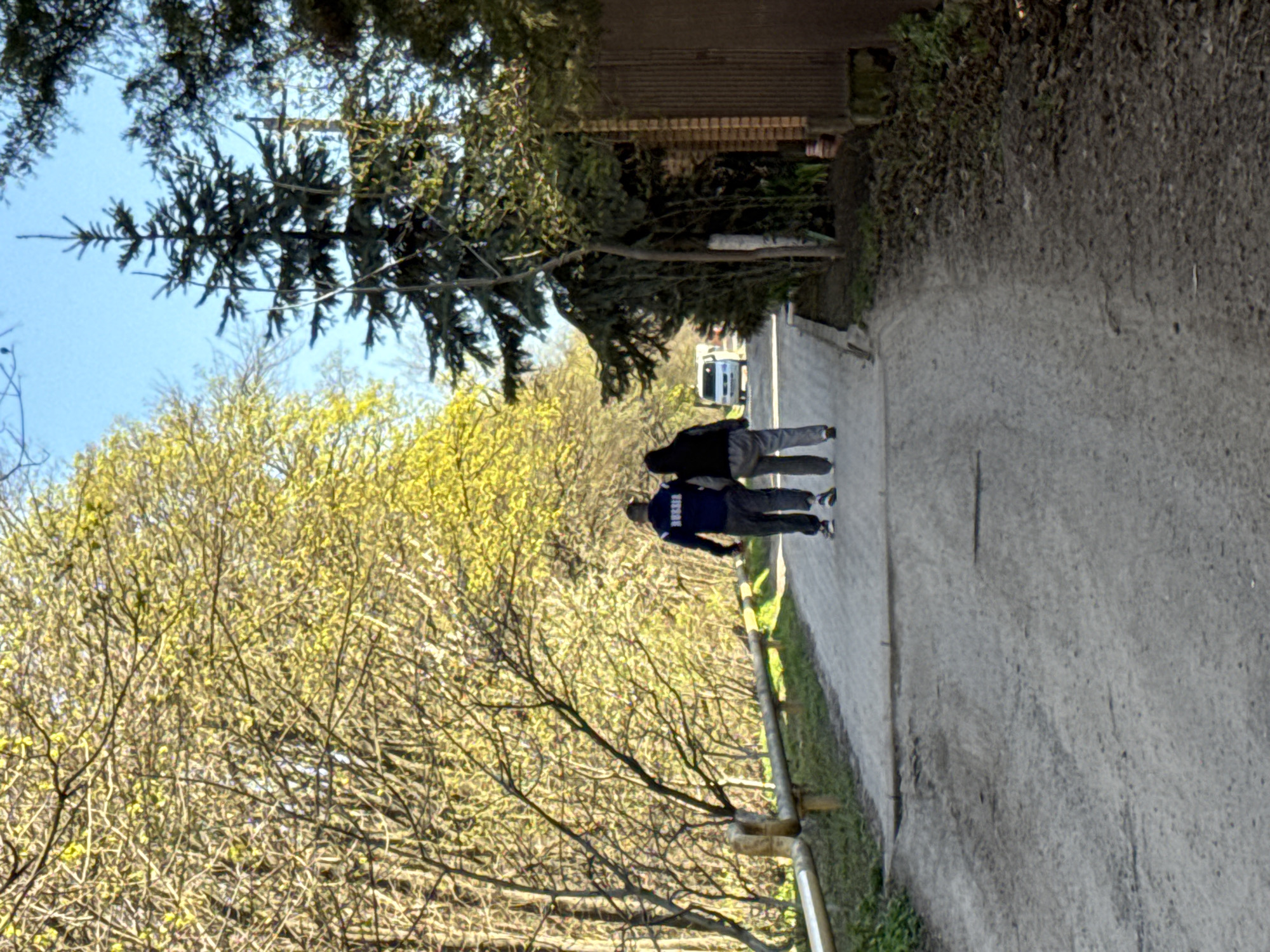 Two individuals are walking along a paved path. One person wears a dark jacket and gray pants, while the other wears a blue jacket and dark pants. Both individuals appear to be walking in the same direction. A white van is visible in the distance. The path is lined with trees. The location is Rostov-on-Don, Russia.