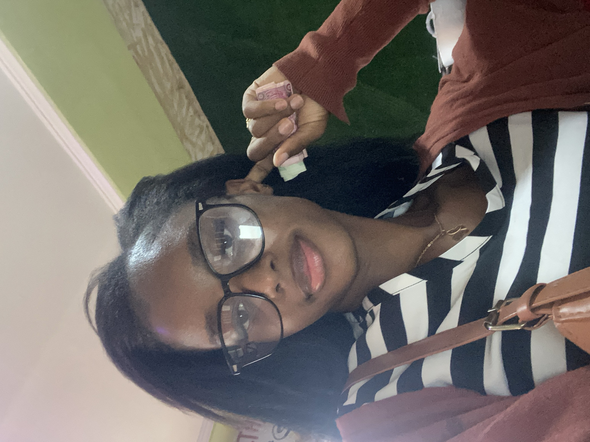 A young Black woman with dark skin, dark hair, and glasses is shown from a low angle. She is wearing a black and white striped shirt, a brown cardigan, and a brown purse. She is holding a wad of pink and green bills in her right hand. The background appears to be an indoor setting with a light green wall and a darker green wall. There is a sliver of a white ceiling visible at the top of the image. Based on visual cues, such as the colors of the currency and the overall ambiance, this image was likely taken in Jos, Nigeria.