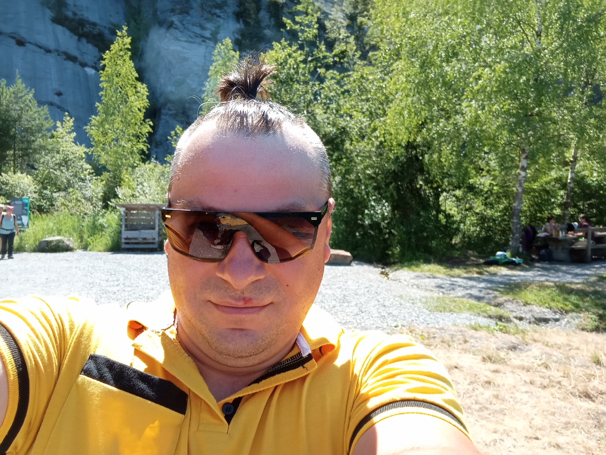 In Emmetten, Switzerland, a man is taking a selfie. He has his hair tied up in a bun and is wearing sunglasses and a yellow polo shirt. The background consists of lush green trees, a rocky cliff face, and a gravel path. In the distance, there are picnic tables and people gathered.