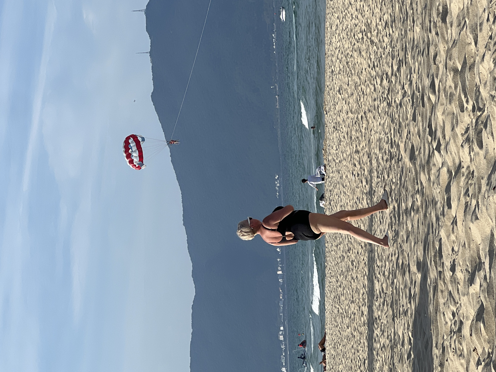 This is a photograph of a beach scene. In the foreground, a blonde woman wearing a black swimsuit and white sunglasses walks across the sand. She is facing away from the camera. The beach is sandy with ripples. In the middle ground, the ocean is visible with some waves and a few people in the water. There is a parasail in the sky above the water, with two people attached to it. In the background, a large, dark, forested mountain range stretches across the horizon. The sky is blue with some scattered clouds. The scene appears to be on a sunny day. The presence of parasailing suggests a tourist beach area, and the mountainous backdrop is characteristic of areas like Sơn Trà Peninsula in Đà Nẵng, Vietnam, which offers such scenic views and recreational activities.