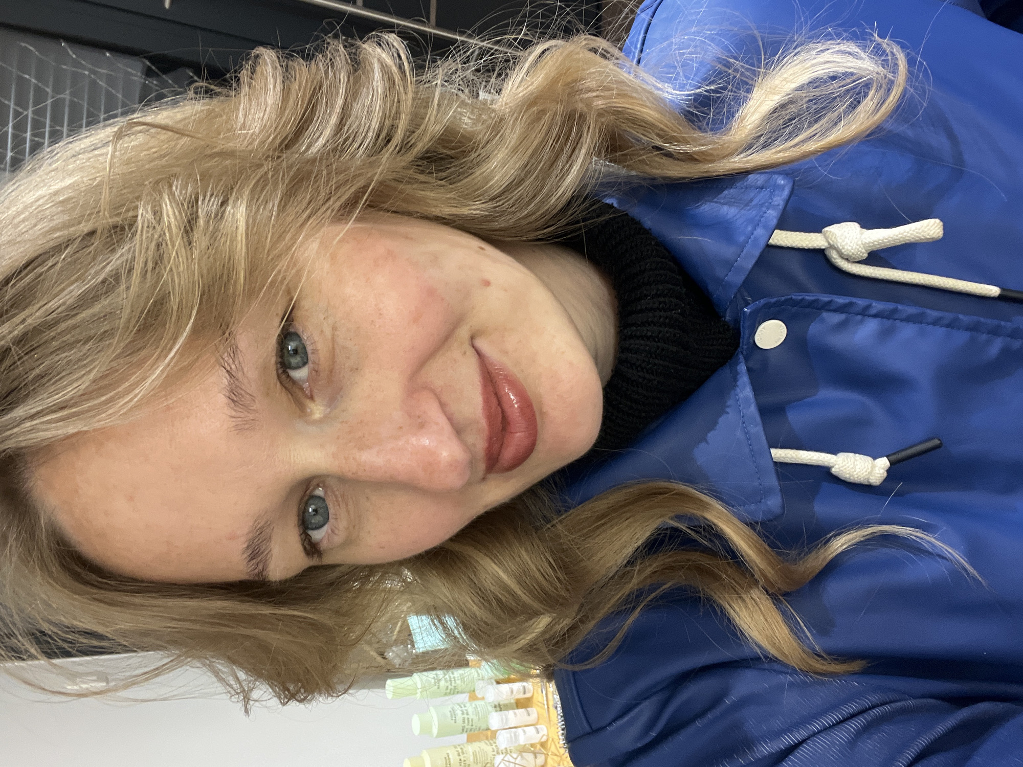 A fair-skinned female subject with blonde, wavy hair, blue eyes, and freckles on her face is depicted in a close-up, chest-up shot. Her lips are closed and coated with a glossy, reddish-brown substance. She is looking directly towards the viewer with a slight smile. The subject is wearing a blue collared jacket with a white drawstring visible at the neckline, and a black ribbed turtleneck garment underneath. The background is out of focus, revealing an indoor setting. To the left of the subject, several light green bottles and small white vials are arranged on a surface, suggesting a retail or salon environment. The scene takes place in the city of Budva, Montenegro.