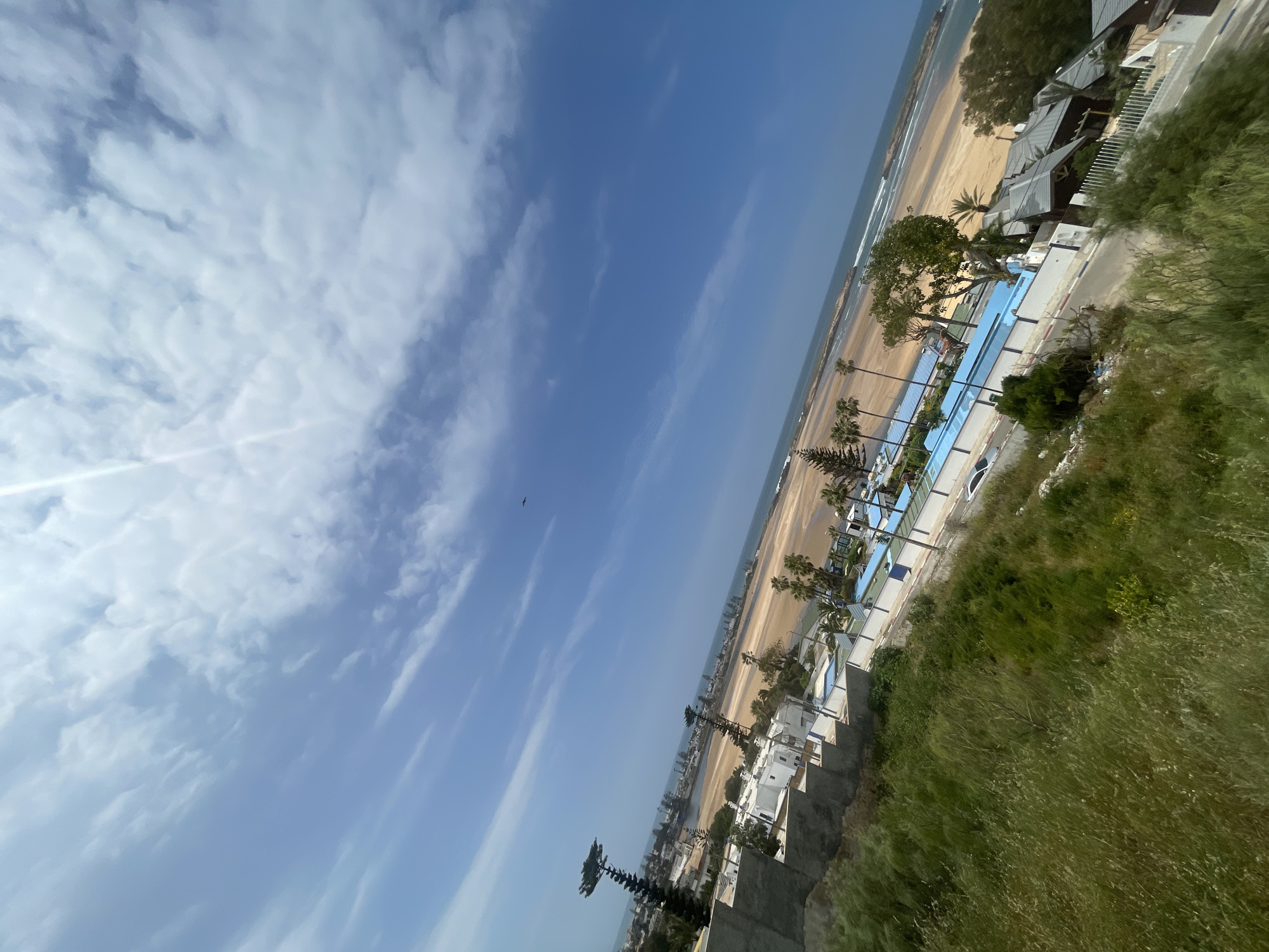 The image captures a vibrant coastal landscape in Oualidia, Morocco, seen from an elevated perspective under a clear, sunny sky. The original image is tilted, presenting the horizon diagonally; when properly oriented, the scene unfolds from a high viewpoint looking down towards the ocean.
The upper portion of the frame is dominated by a bright blue sky, adorned with scattered white clouds ranging from wispy cirrus to more textured, patchy formations. A bright sun flare is visible in the upper left quadrant.
Below the sky, the vast expanse of the Atlantic Ocean meets a wide, sandy beach. Gentle waves can be seen breaking on the shore, and the sand appears light brown. Along the coastline, a series of white-walled buildings with dark sloped roofs are nestled, possibly indicating hotels or residences. Adjacent to some of these structures, a long, bright blue swimming pool is prominently visible, running parallel to the beach.
Numerous palm trees and other green trees are interspersed among the buildings and along the beach, adding to the lush coastal feel. A paved road or pathway winds through the developed area, and a white car is partially visible on it. In the foreground, particularly on the right side, a wild, overgrown hillside covered in vibrant green grass and shrubs provides a natural contrast to the built environment. A small, dark bird is captured in flight against the blue sky, adding a subtle touch of life to the expansive view.
The scene depicts a peaceful and bright day, likely morning or early afternoon, with no specific human activity clearly discernible beyond the presence of the coastal development. No visible text is present in the image.